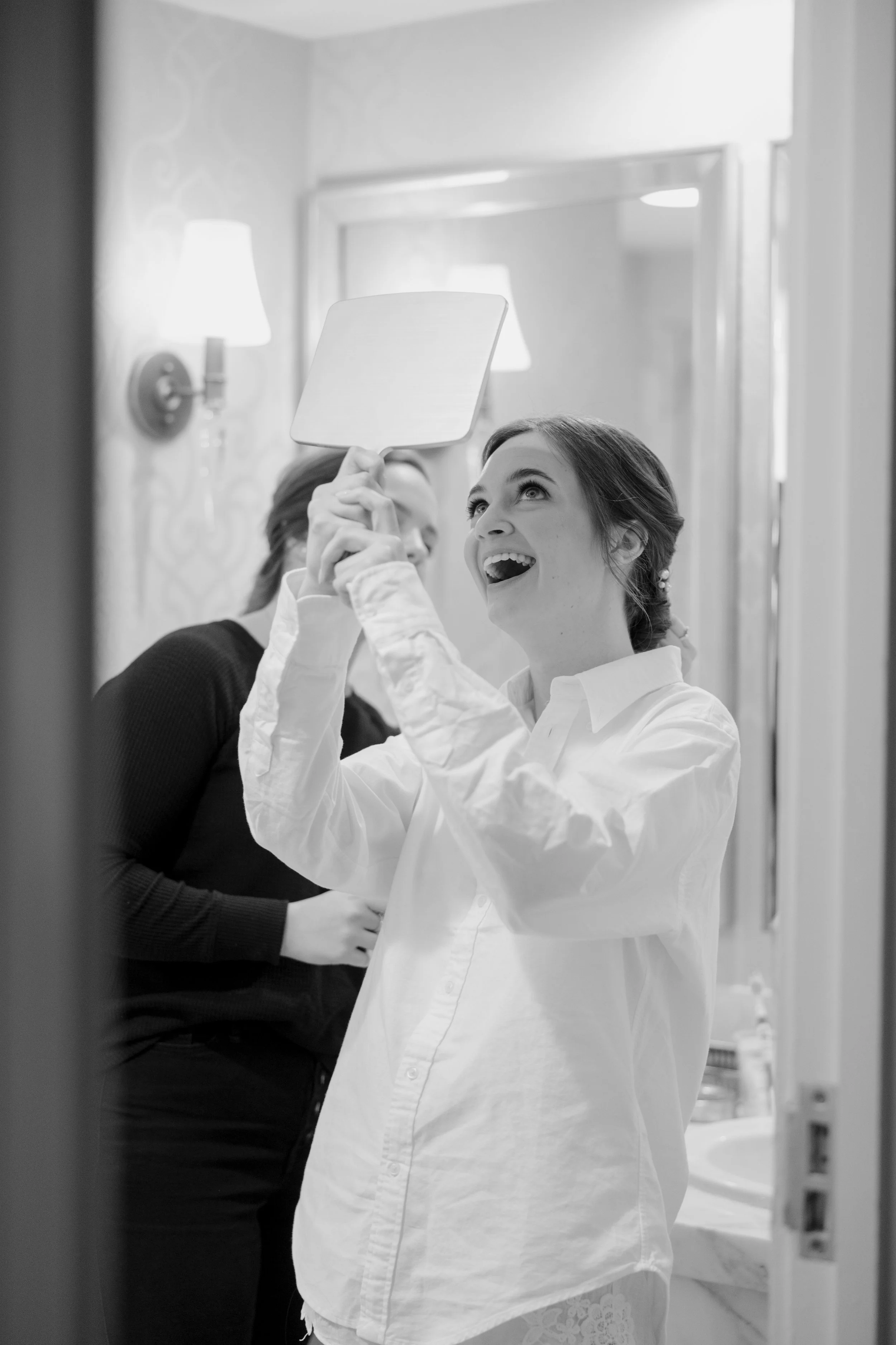 A woman with styled hair and earrings, wearing a white blouse, looking happy as she holds a mirror, with a person in a dark sweater behind her in a mirror reflection, in a well-lit room with lamps and bathroom fixtures.