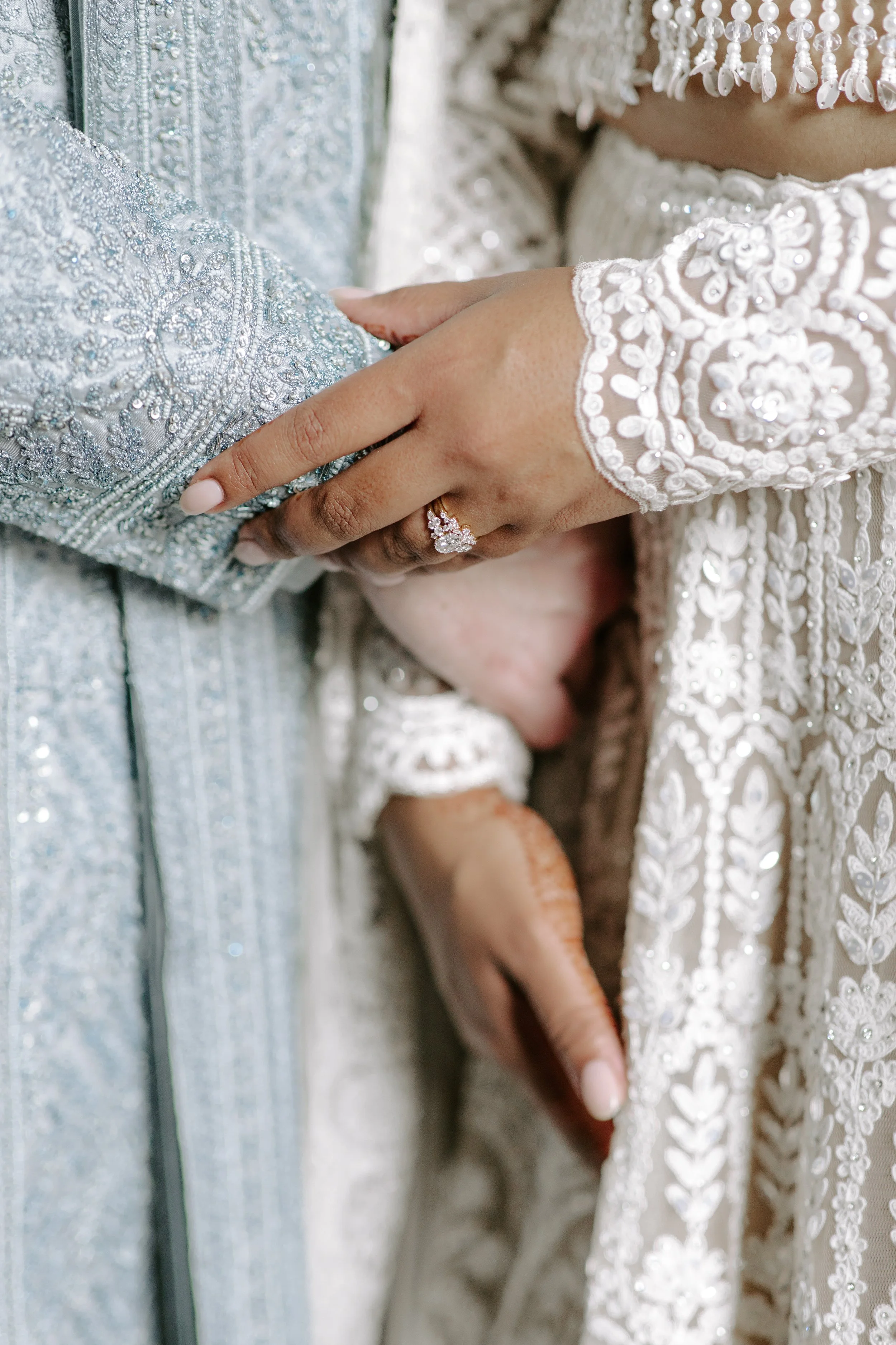 Close-up of a bride and groom holding hands, highlighting the bride's engagement ring and wedding dress with intricate embroidery and beadwork, and the groom's silver metallic suit.