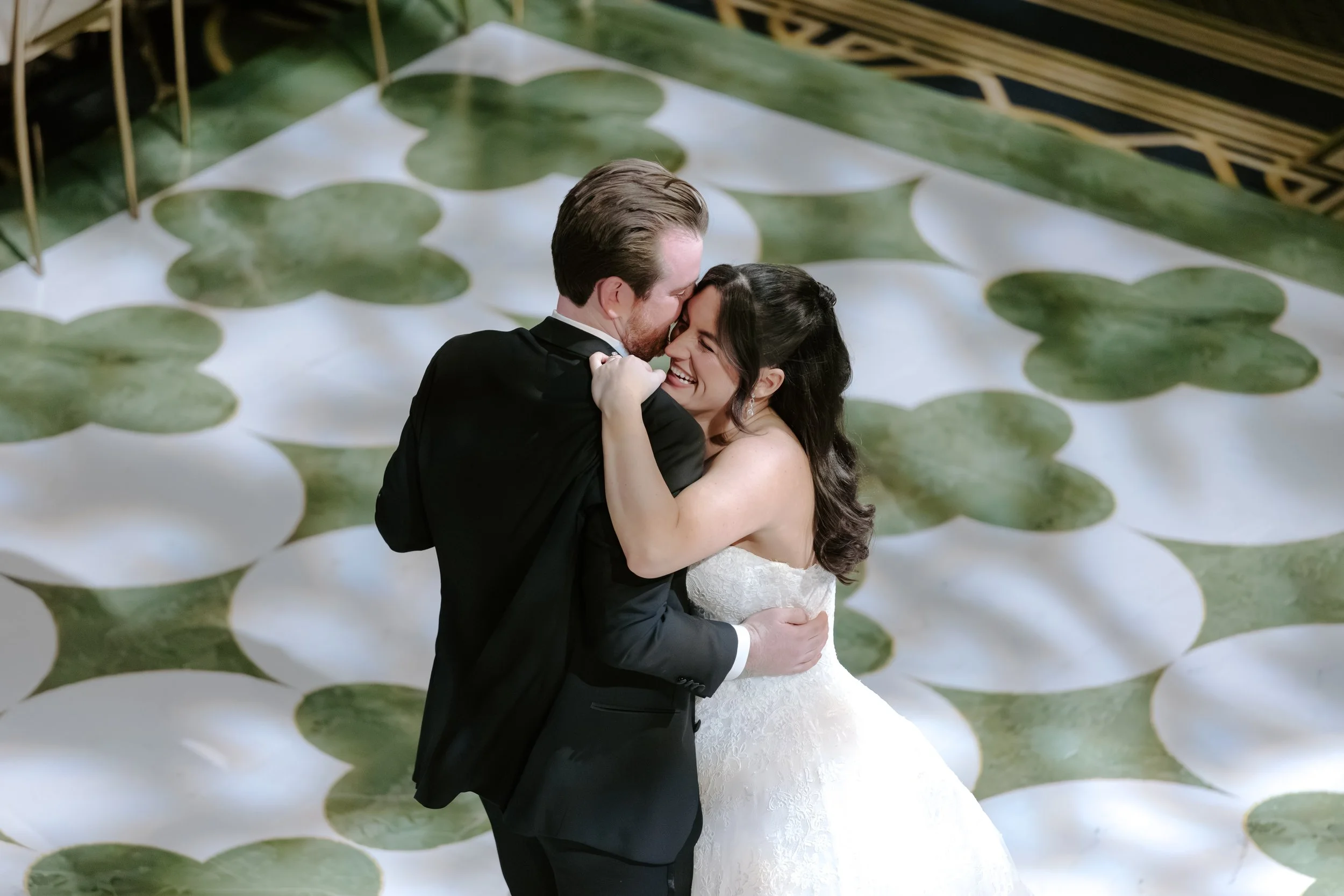 A bride and groom sharing a dance, with the groom in a black tuxedo and the bride in a white wedding dress. They are smiling and laughing, embracing each other, on a floral-patterned dance floor.