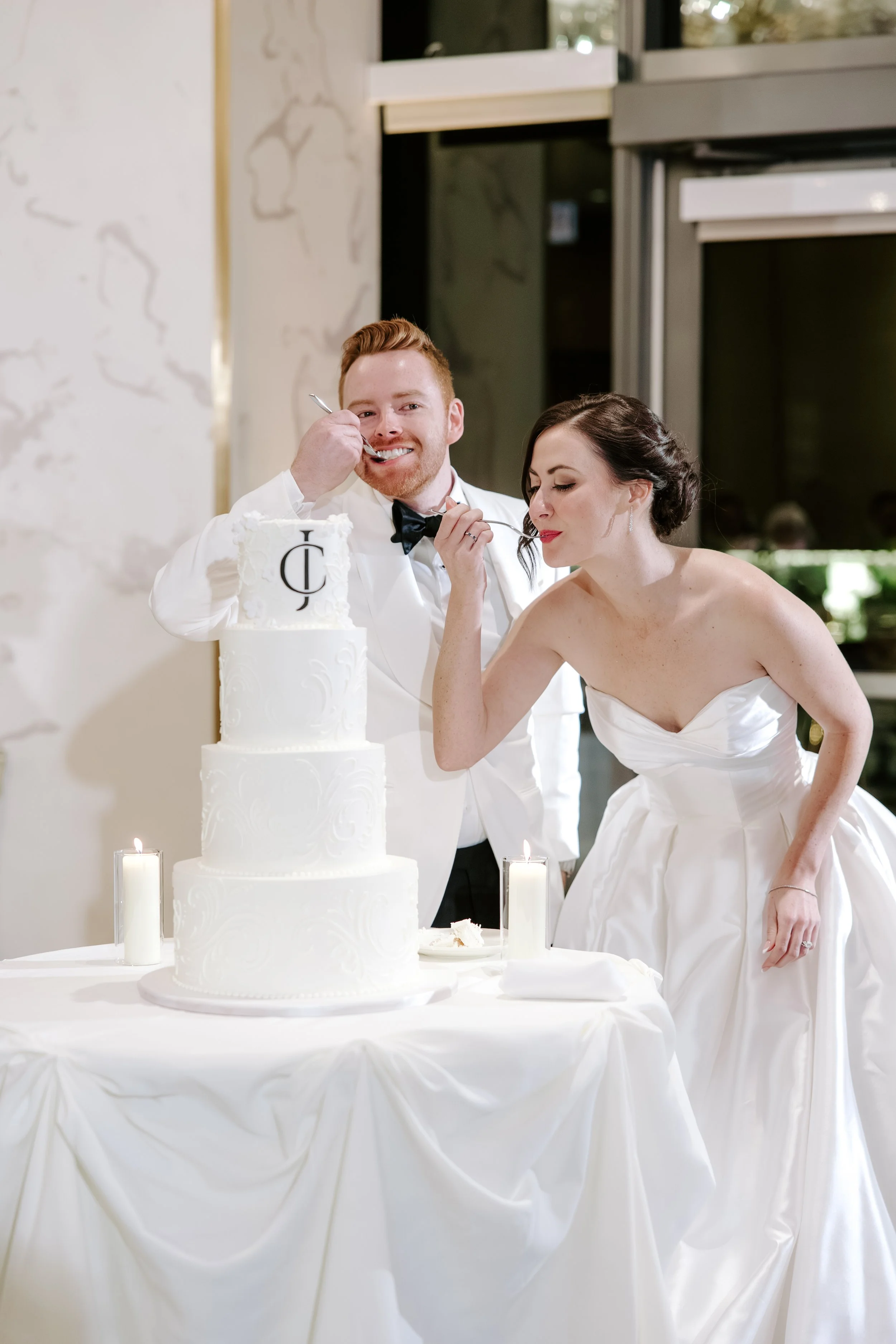 A bride and groom in wedding attire sharing a piece of cake at their wedding reception. The groom is smiling as he feeds the bride, and the bride has her eyes closed, about to taste the cake. A white wedding cake with monogram decoration and lit cand