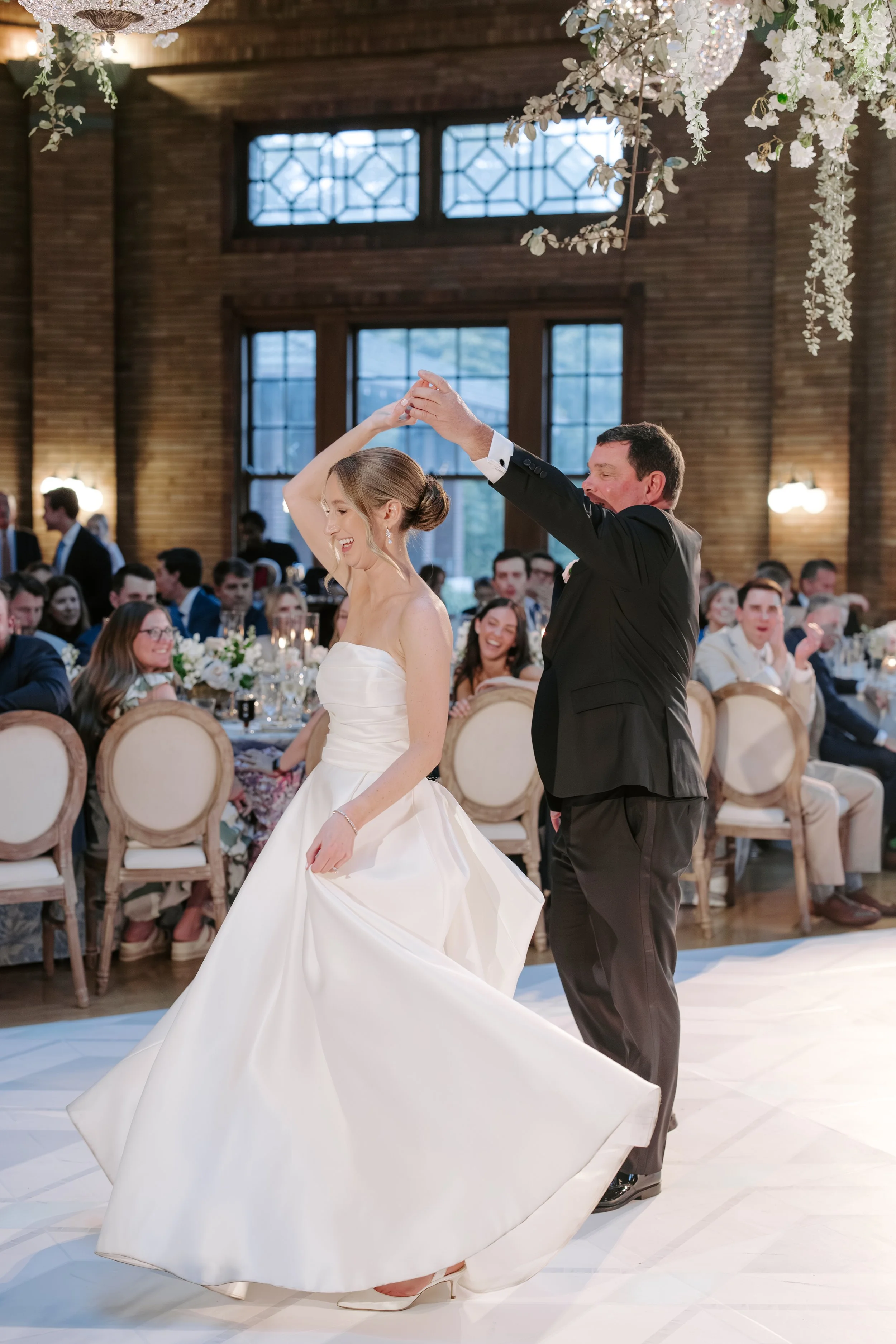 A bride and groom are dancing at their wedding reception indoors, with guests sitting at tables in the background. The bride wears a strapless white wedding gown, and the groom wears a black tuxedo. The bride is smiling and has her hair styled in an 