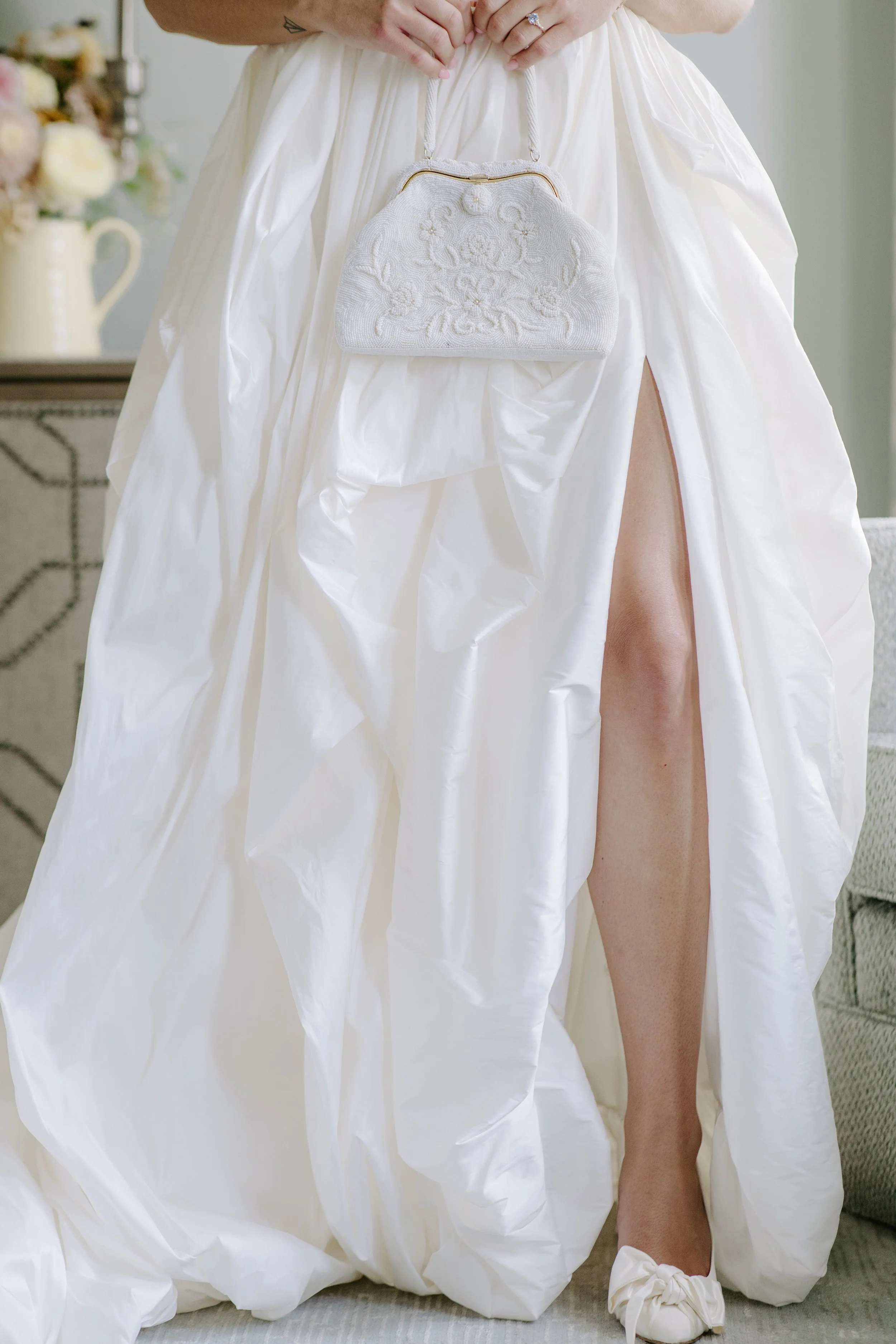 A bride wearing a white dress is kneeling, showing her leg through a slit in the dress. She holds a white, embroidered handbag. The setting appears to be indoors with some blurred flowers in the background.