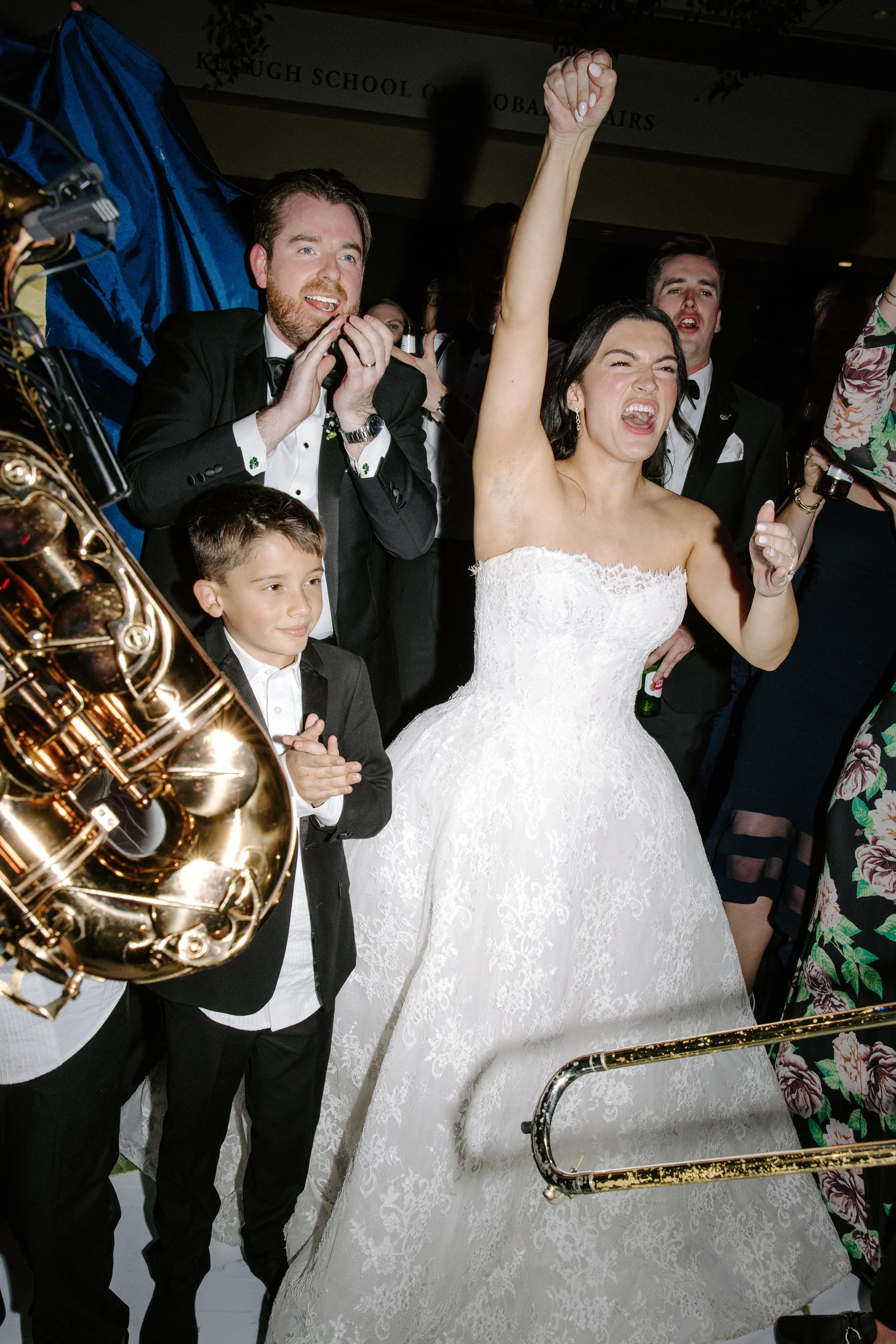A bride in a white lace wedding dress celebrating with a group of people, including a woman with dark hair and a woman with glasses, at a wedding reception with musical instruments and a banner in the background.