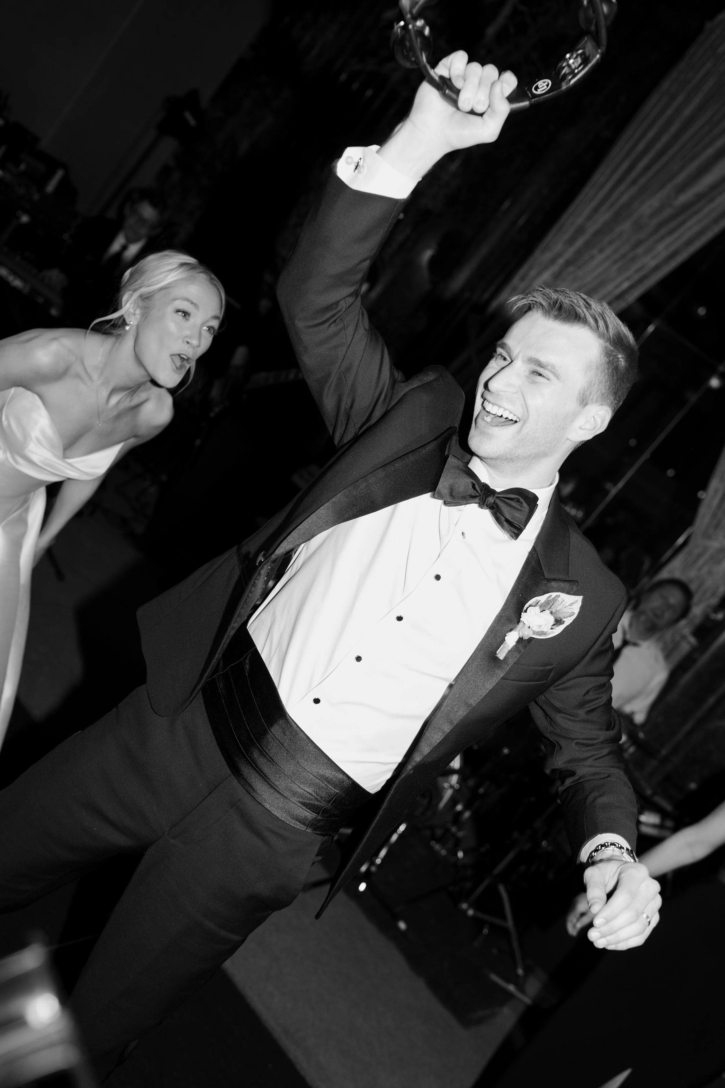 A man in formal attire holding a circular object above his head and smiling, with a woman in a strapless dress looking surprised in the background, at an indoor event.