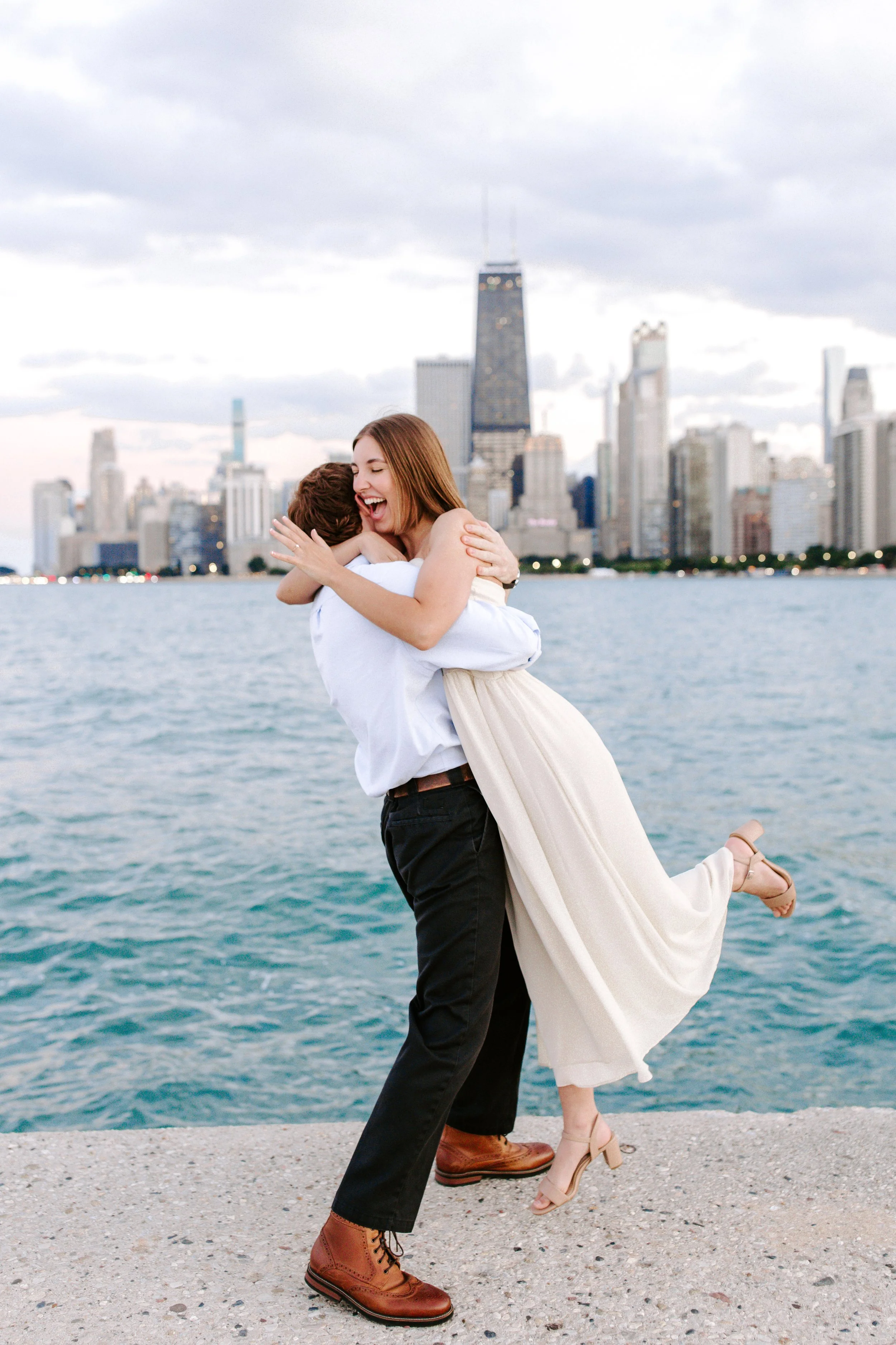 A couple hugging by the water with a city skyline in the background, the woman wearing a strapless dress and high heels, and the man in dark pants, dress shoes, and a long-sleeve shirt.