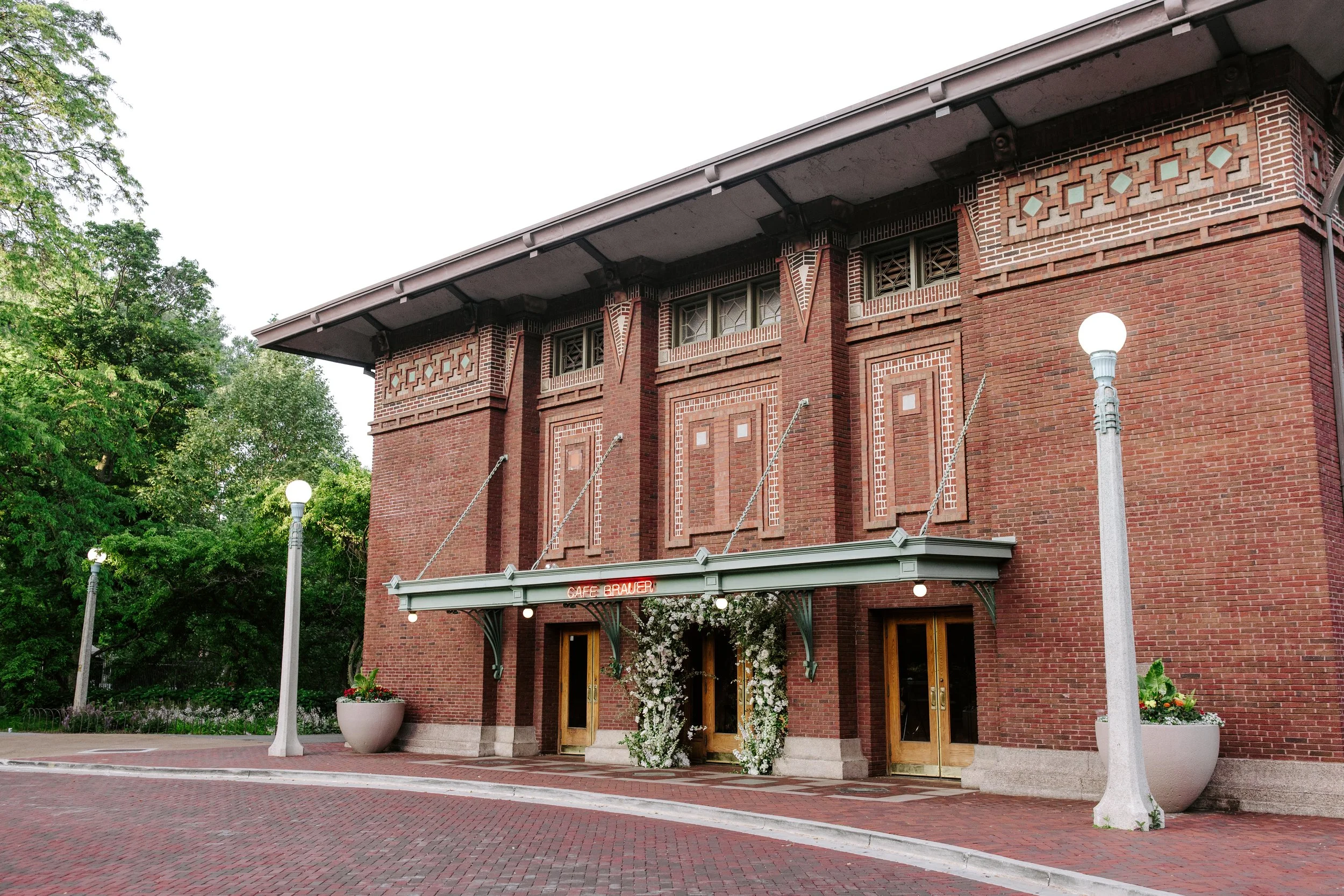 Red-brick building with decorative brickwork and a green awning over the entrance, flanked by white street lamps and pots with flowers, surrounded by green trees.