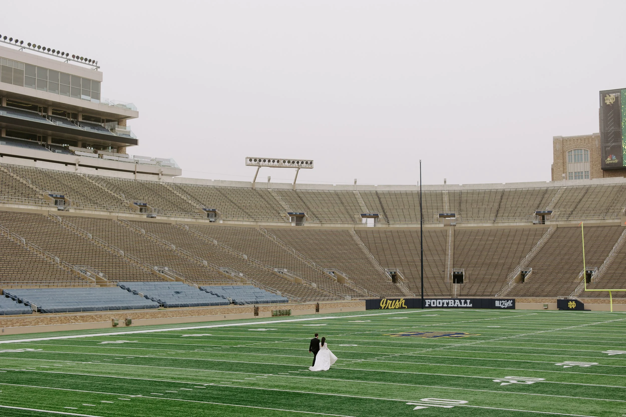 A bride and groom walking on a football field in a large stadium, with empty seats and a cloudy sky overhead.