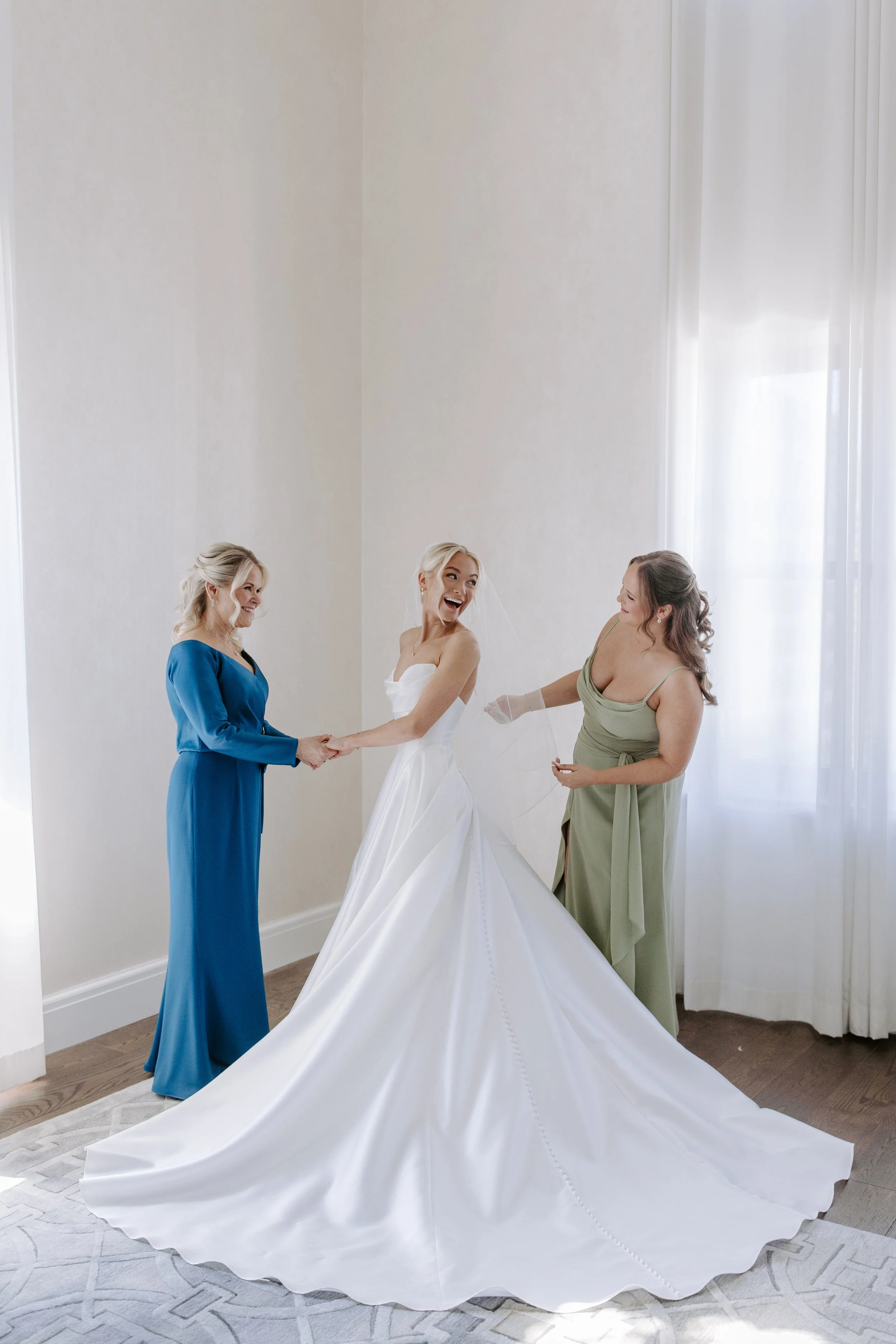 A bride in a white wedding dress holding hands with a woman in a blue dress, while another woman in a green dress stands nearby, smiling, in a brightly lit room with white walls and large windows.
