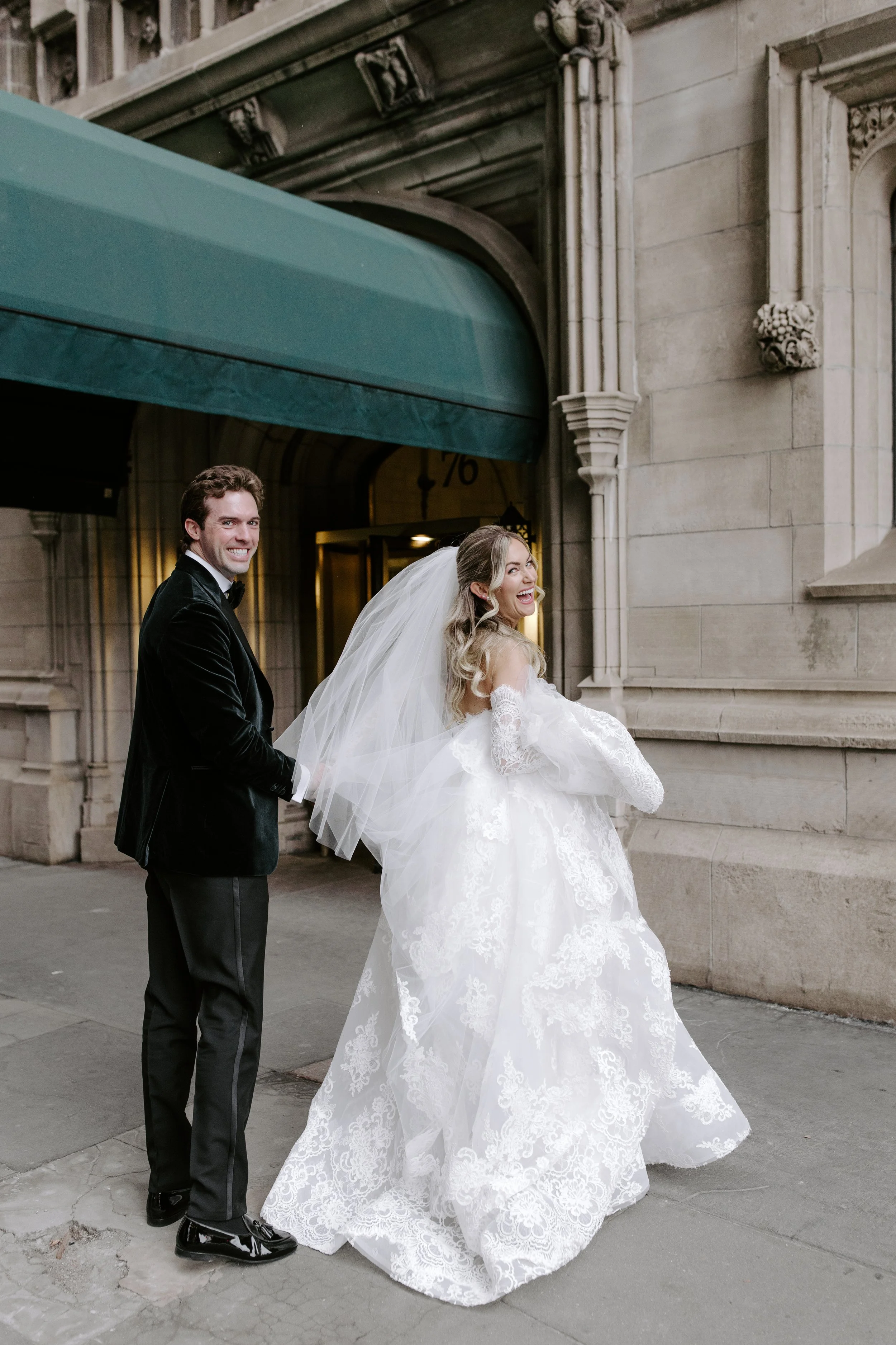 A smiling bride in a white lace wedding gown and veil holding the groom's hand from behind, standing on a city street in front of an ornate stone building, with a smiling groom in a black tuxedo.