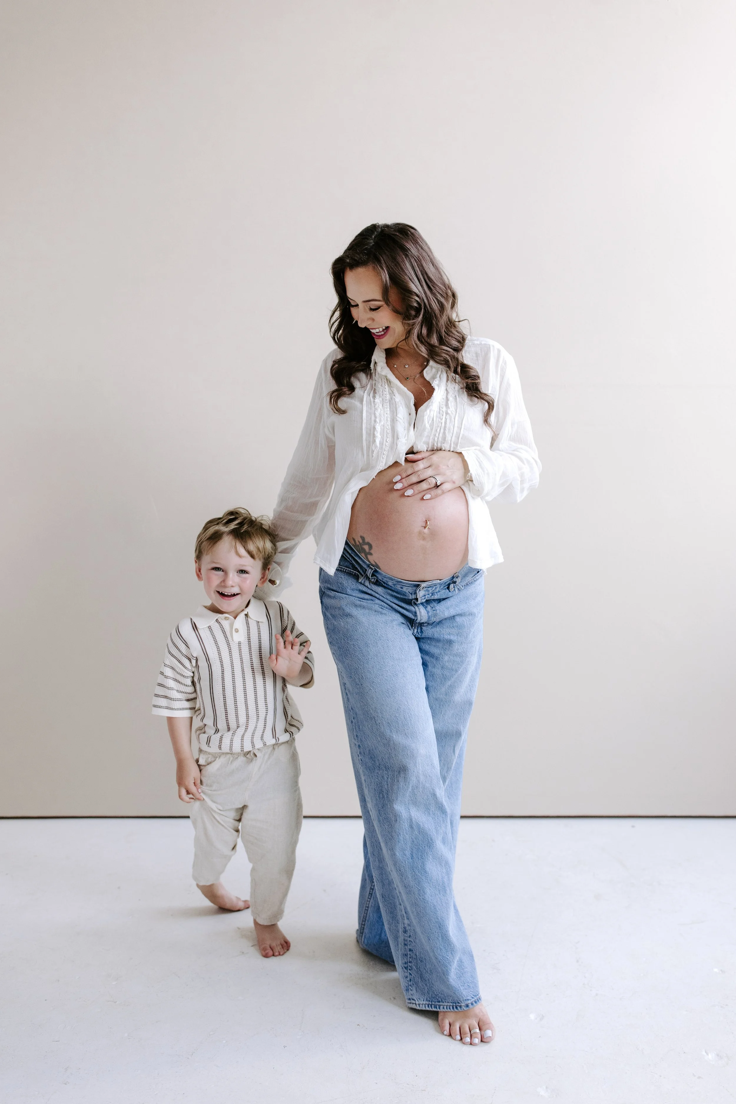 A pregnant woman with long wavy brown hair smiling and holding her belly, wearing a white blouse and jeans, standing next to a young boy with curly hair wearing a striped shirt and beige pants, both standing against a plain off-white background.