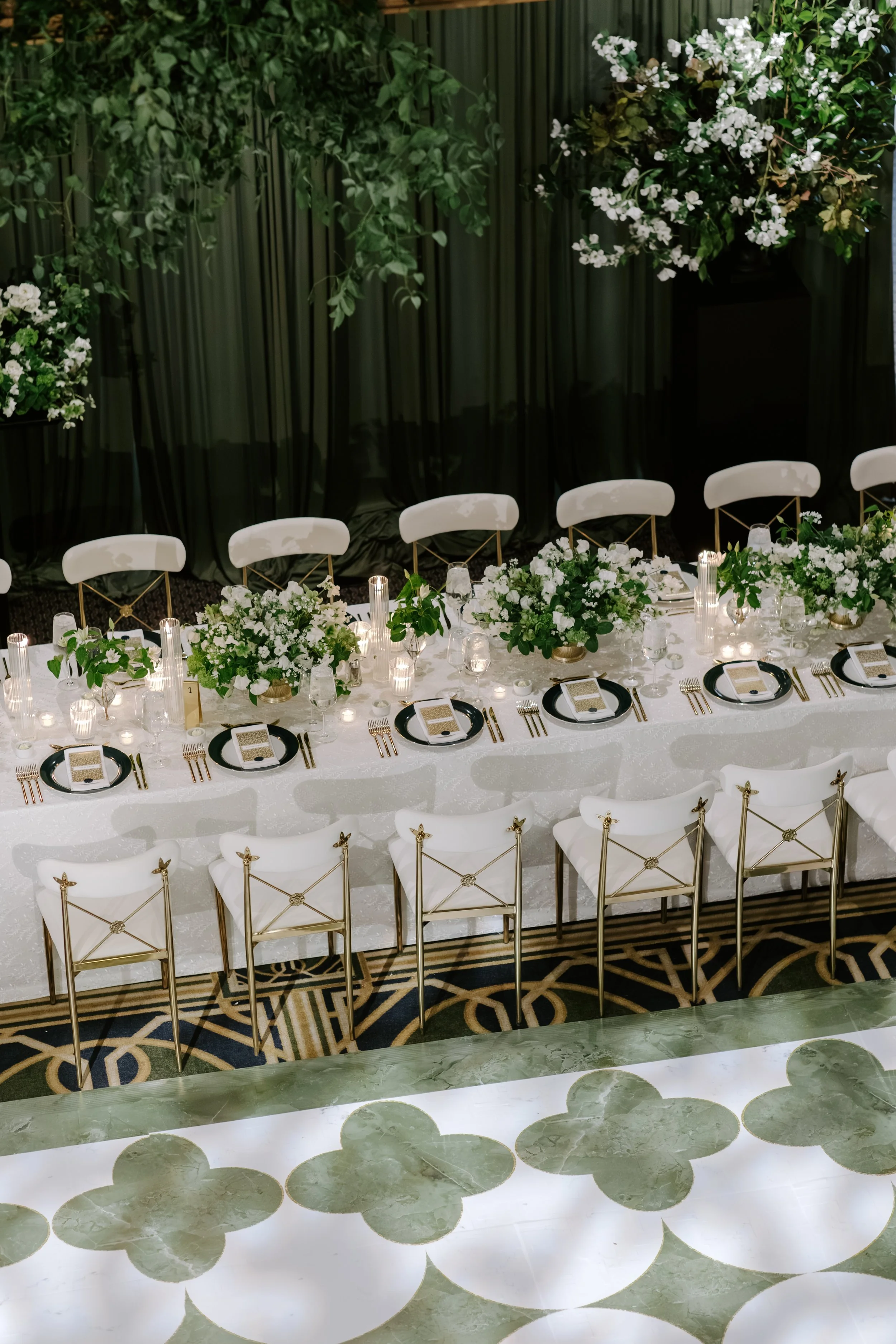Elegant banquet table with white tablecloth, floral centerpieces, black and white plates, gold utensils, and glassware, set against a backdrop of dark curtains and greenery.