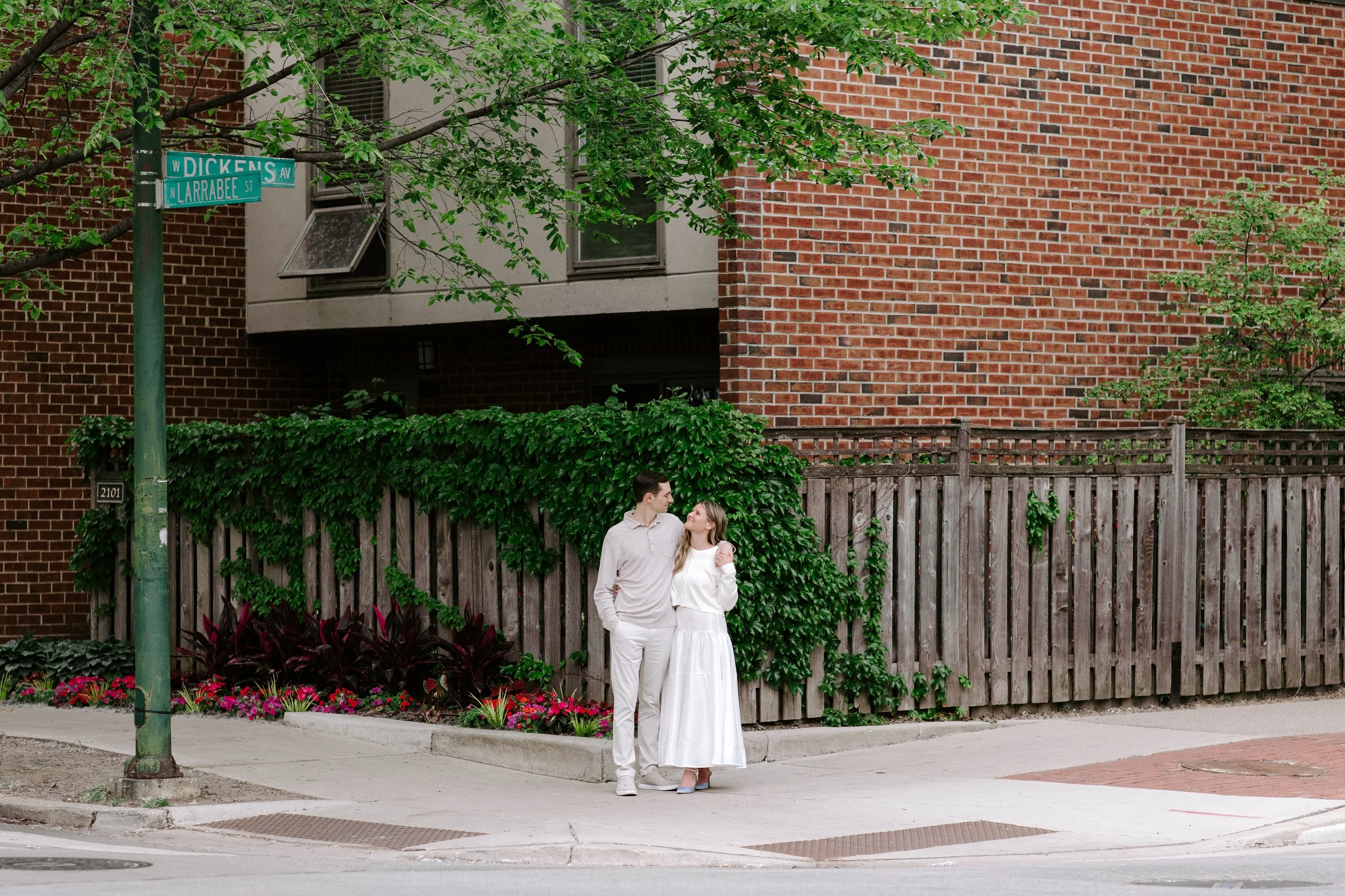A young couple stands close together on a sidewalk next to a wooden fence, with a large tree and colorful flowers in the background. The woman is wearing a white dress and the man is dressed in white pants and a light-colored shirt. They are looking 
