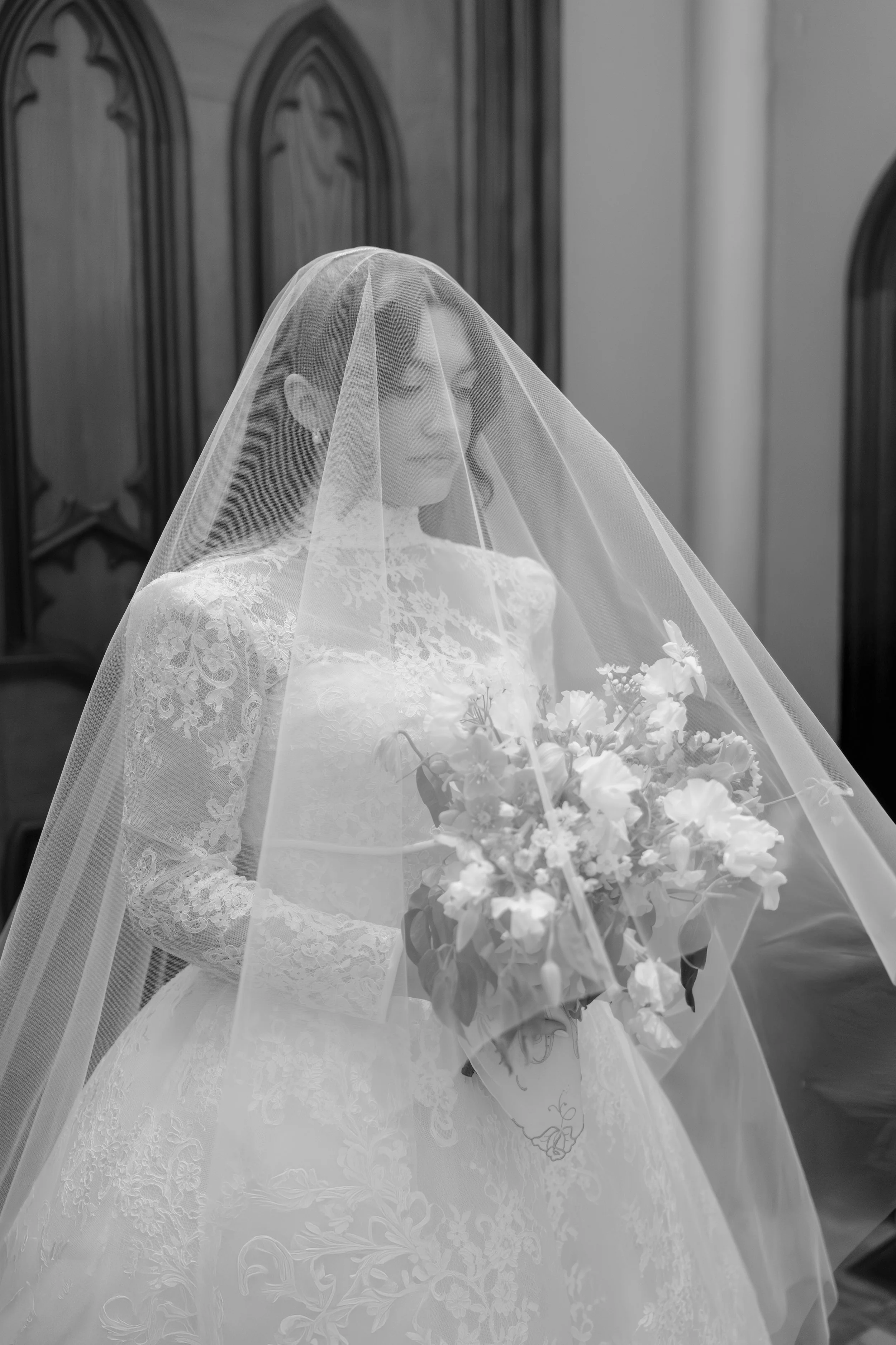 Black and white photo of a bride in a lace wedding dress with a veil, holding a bouquet of flowers, standing indoors near wooden furniture.