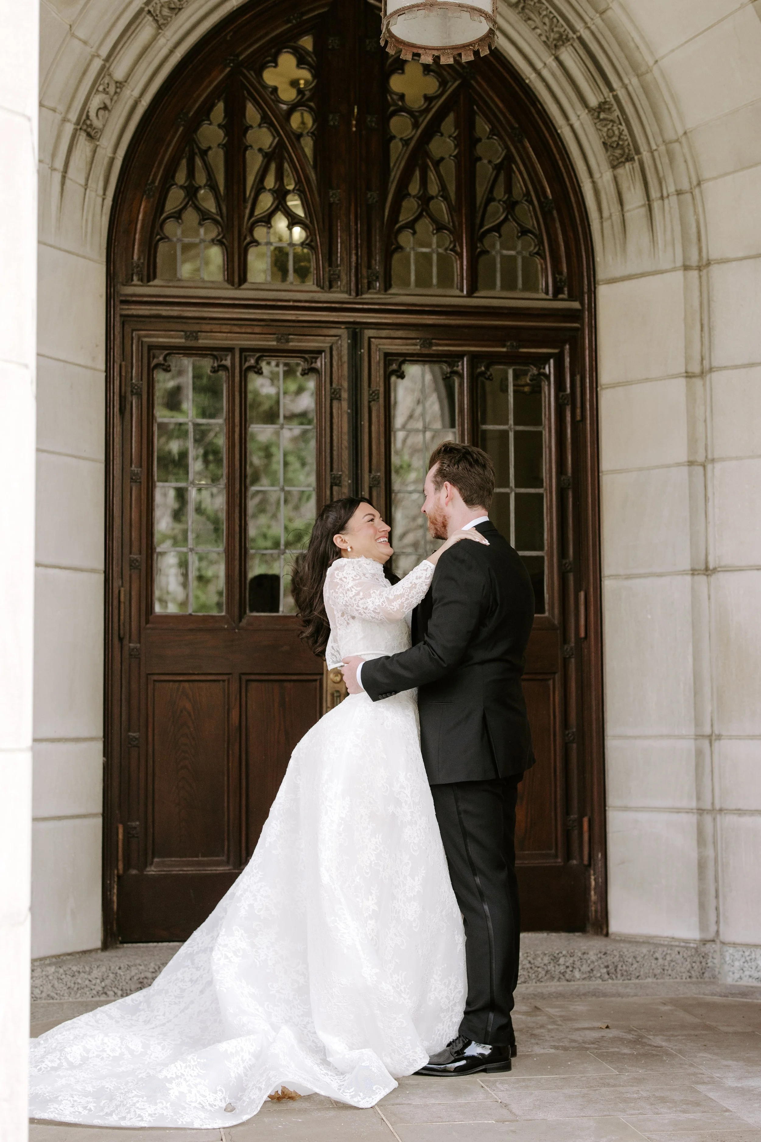A bride and groom sharing a dance in front of a large wooden and glass door at a wedding venue, with the bride wearing a white lace gown and the groom in a black tuxedo.