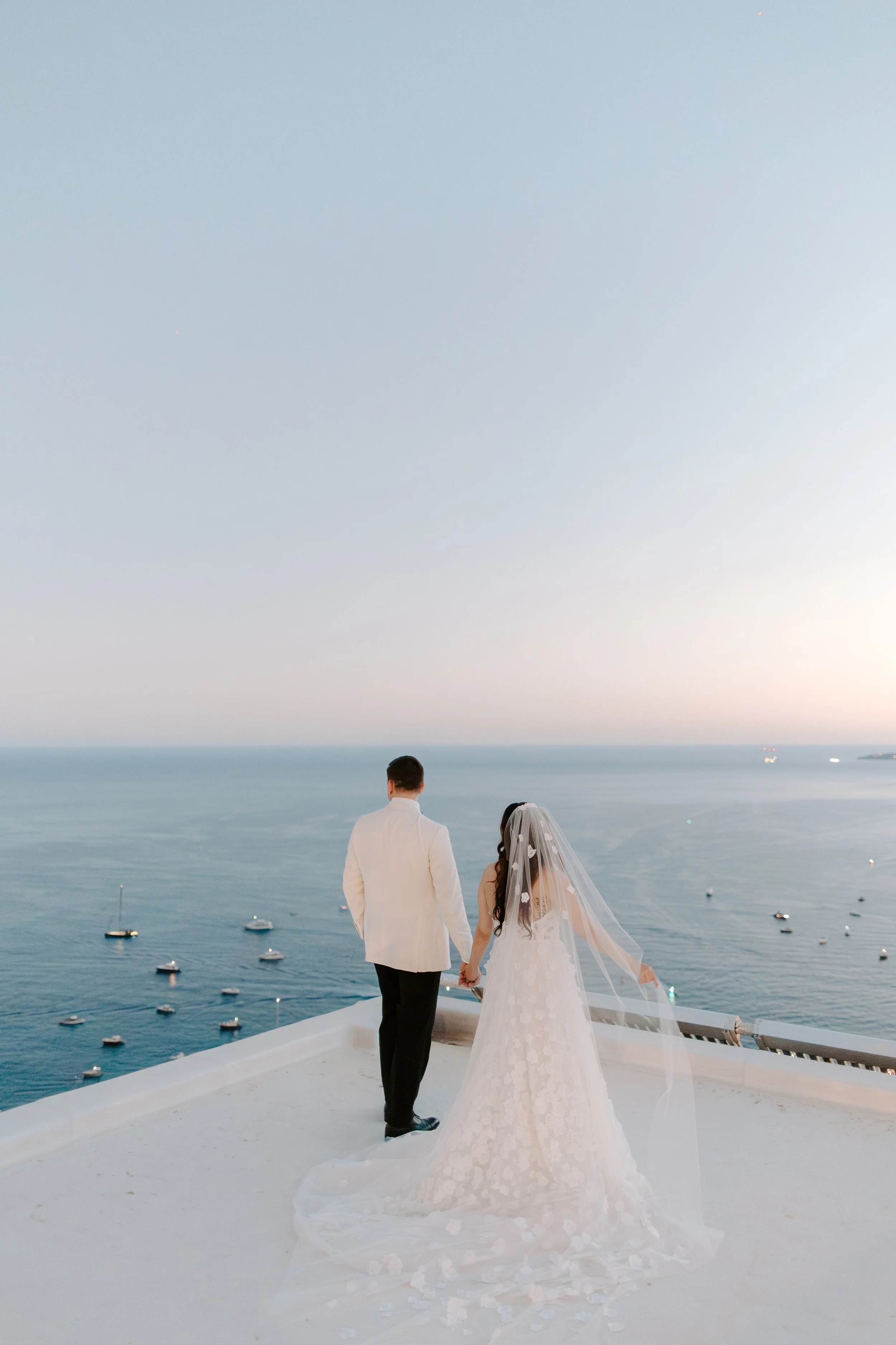 A bride and groom holding hands and walking on a white platform overlooking the ocean, with boats in the water, during sunset.