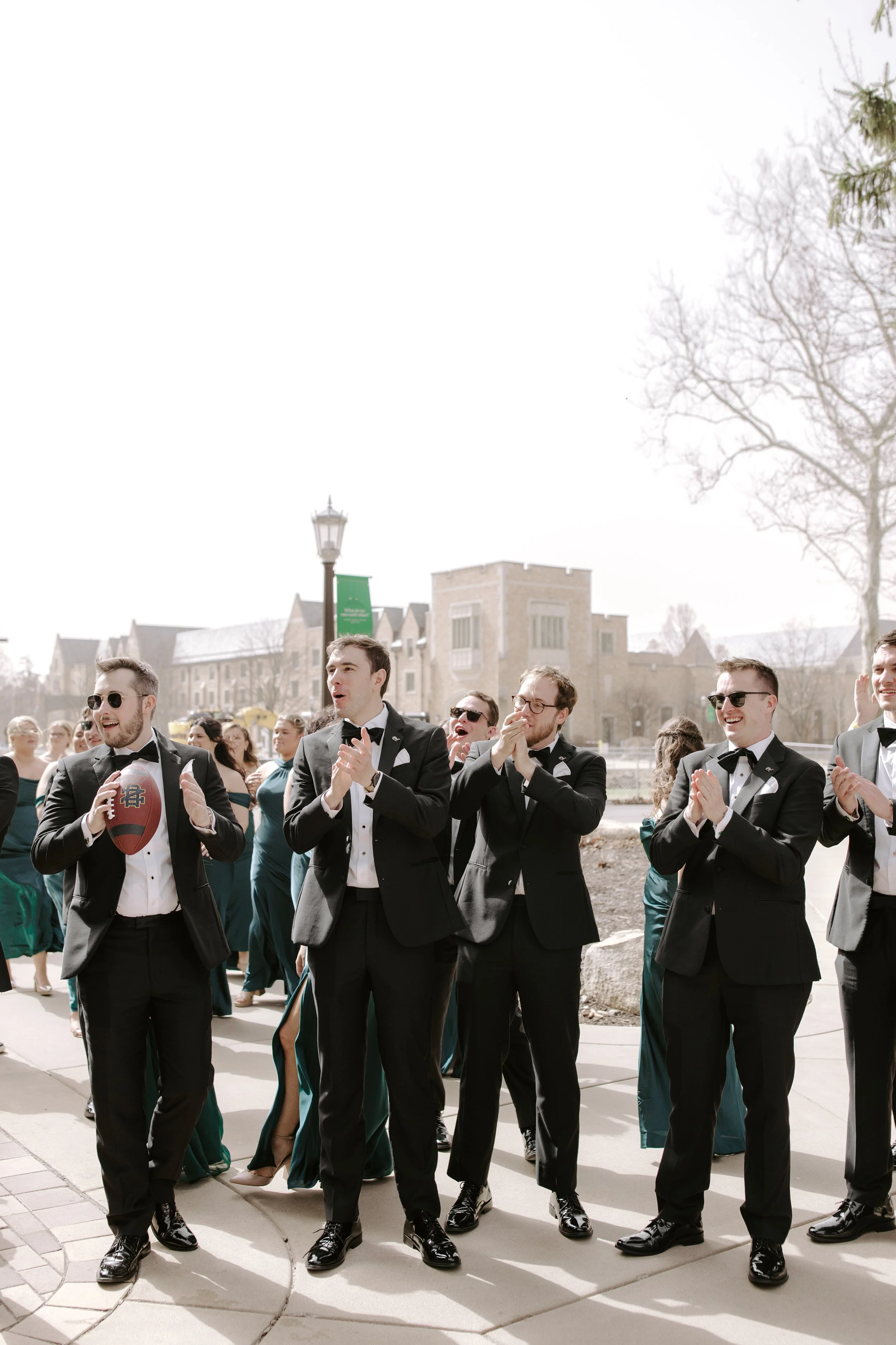 Group of men in tuxedos with green and black pants, some wearing sunglasses, walking outdoors, some clapping, with a crowd and buildings in the background on a sunny day.