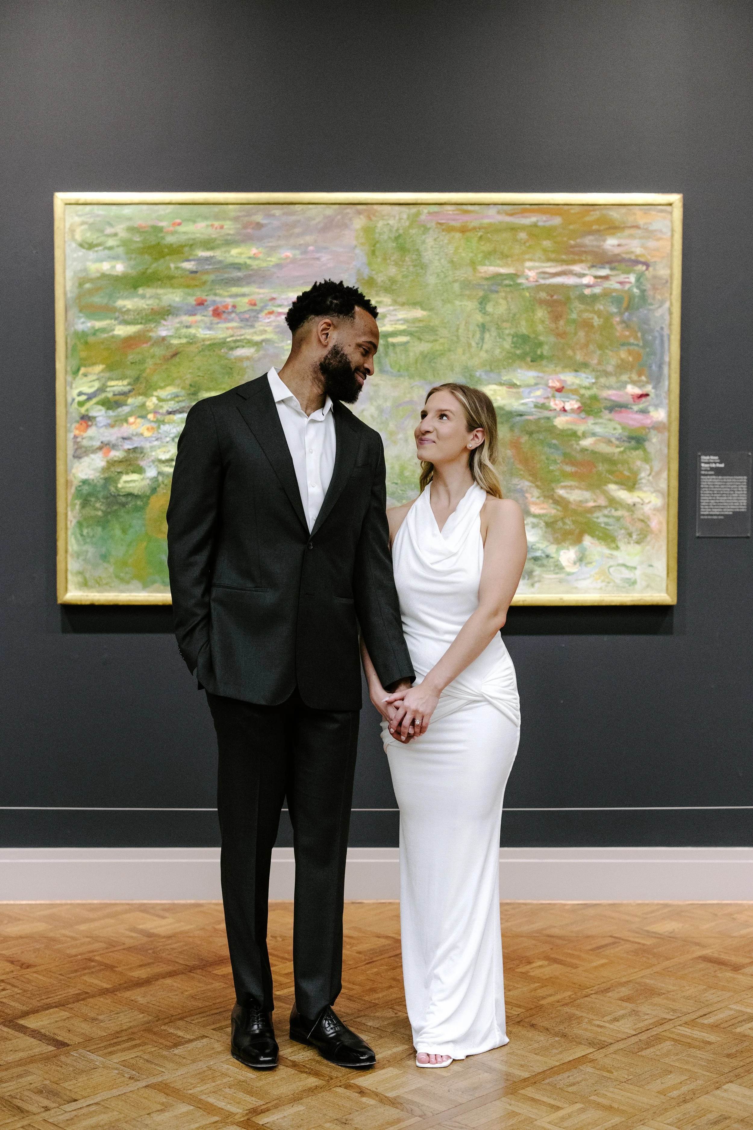 A man and woman holding hands and looking at each other in an art gallery, with a colorful painting of water lilies behind them.