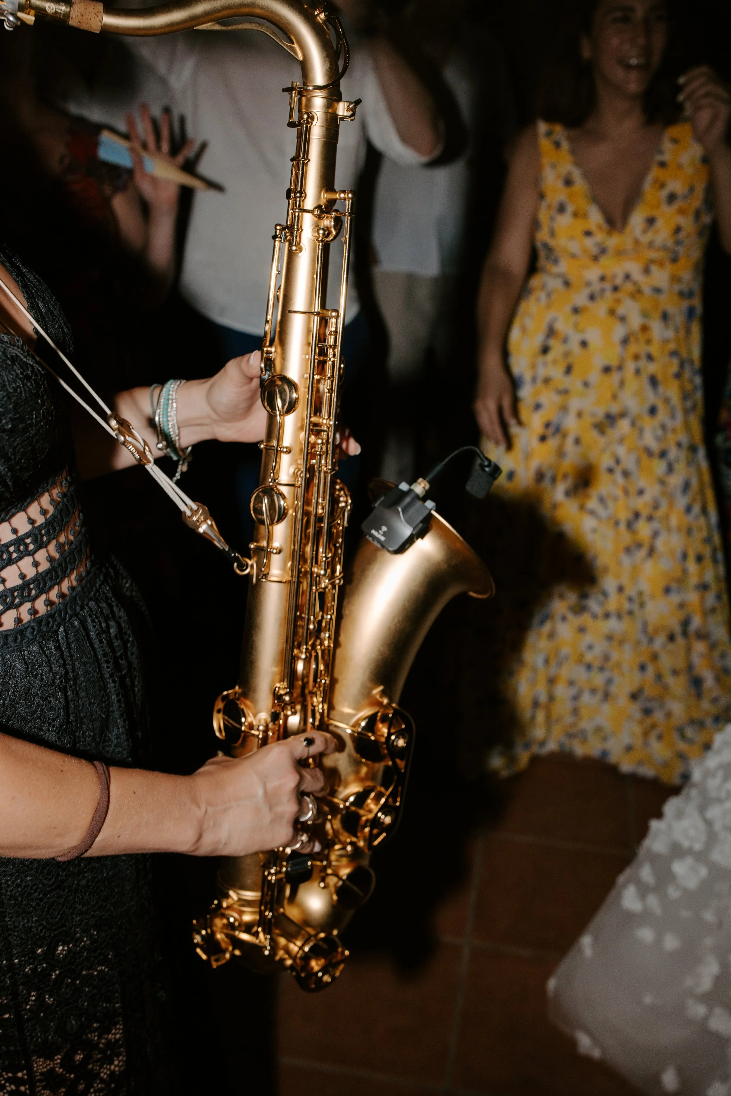 A person holding a gold saxophone at a social gathering, with a woman in a yellow floral dress in the background.