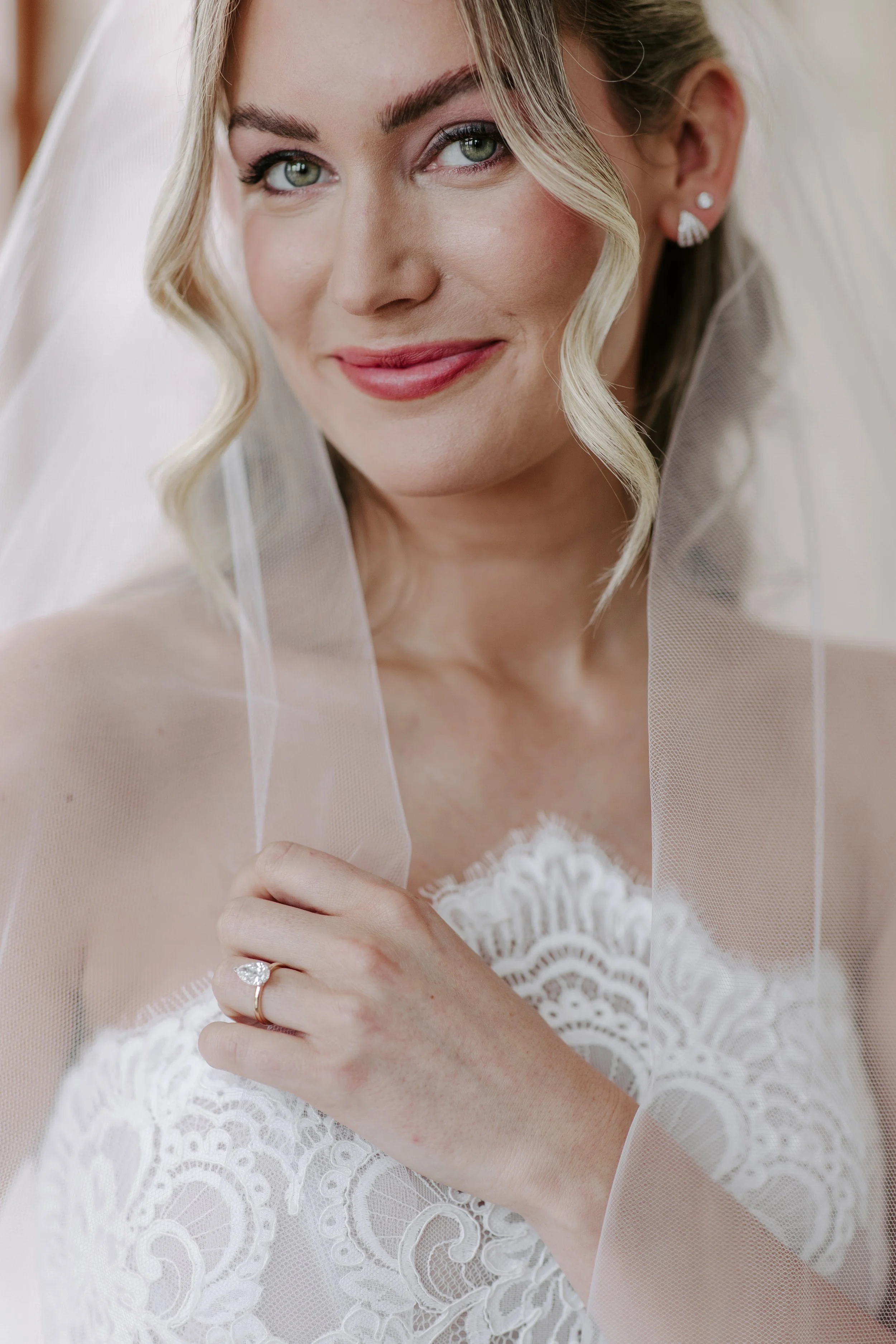 Close-up of a smiling woman on her wedding day, wearing a white lace dress and a sheer veil, with a diamond ring on her finger and pearl earrings.