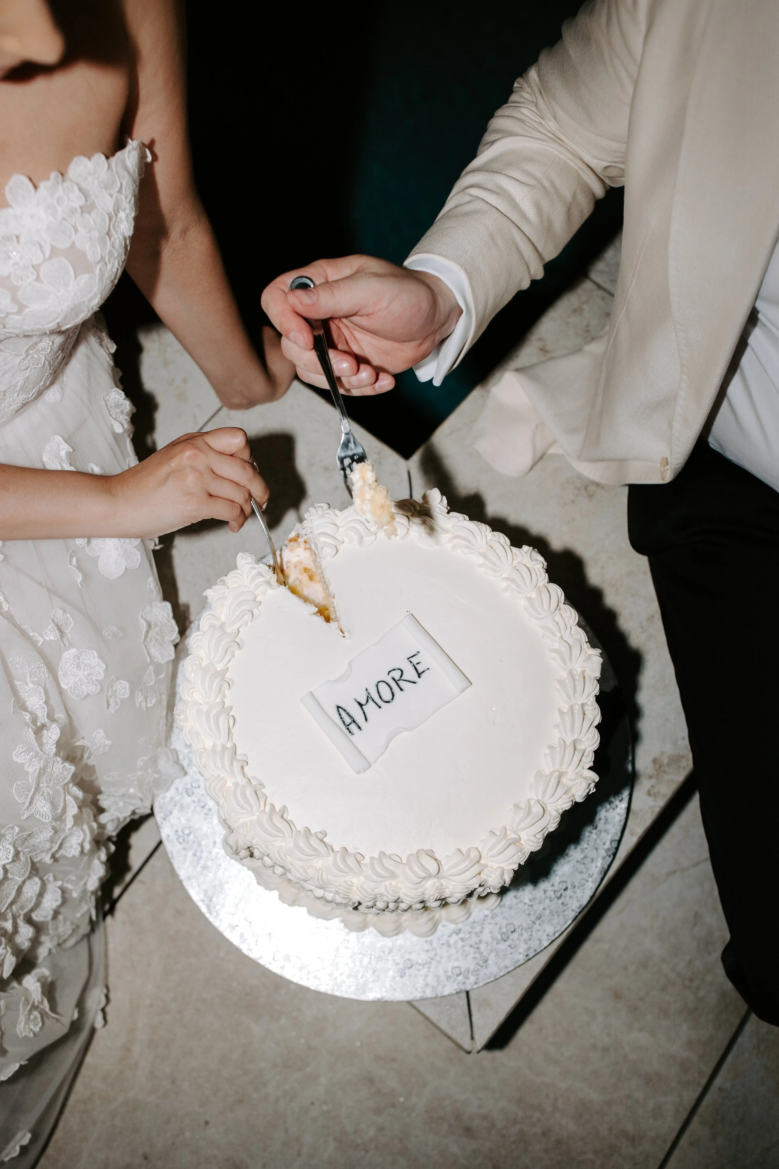 A white wedding cake with a slice being cut, on a silver cake stand. The cake has piped white frosting and a sign that reads 'AMORE'. A bride in a white embroidered dress and a groom in a beige suit are cutting the cake together.