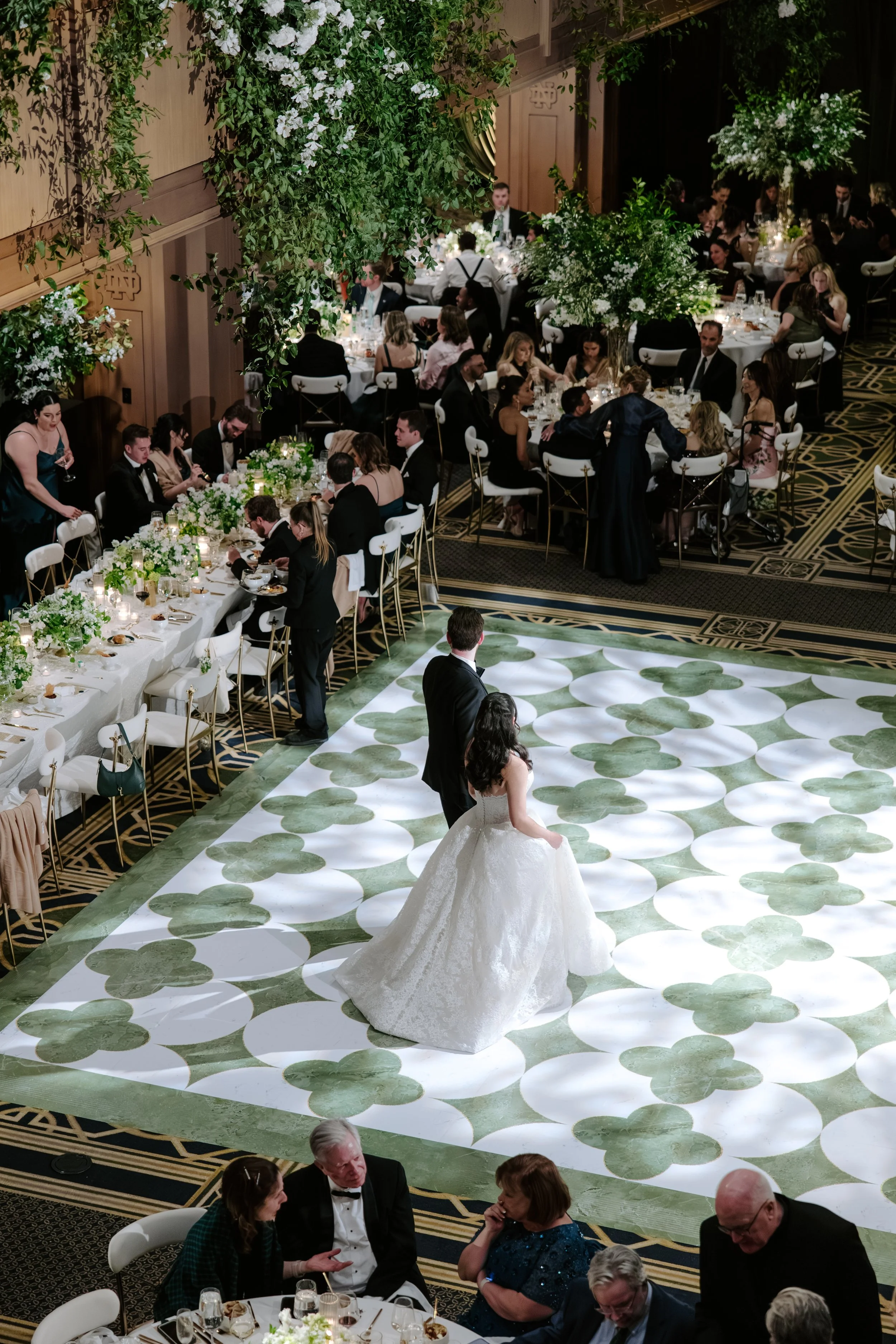 A bride and groom are dancing on a decorated dance floor at a wedding reception, with guests seated at round tables and floral arrangements hanging overhead.
