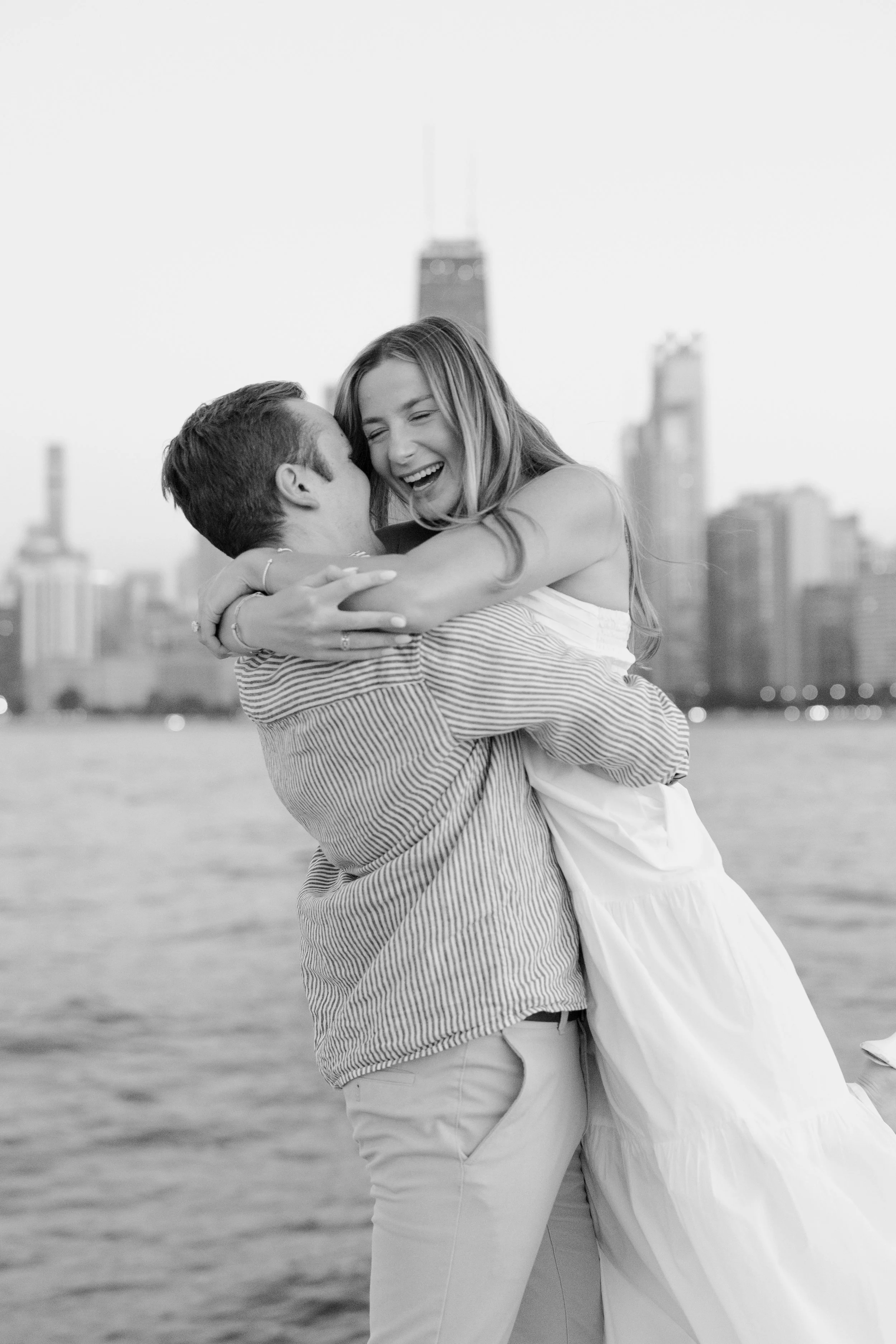 A man lifting and embracing a woman by a body of water, with a city skyline including tall buildings in the background, in black and white.