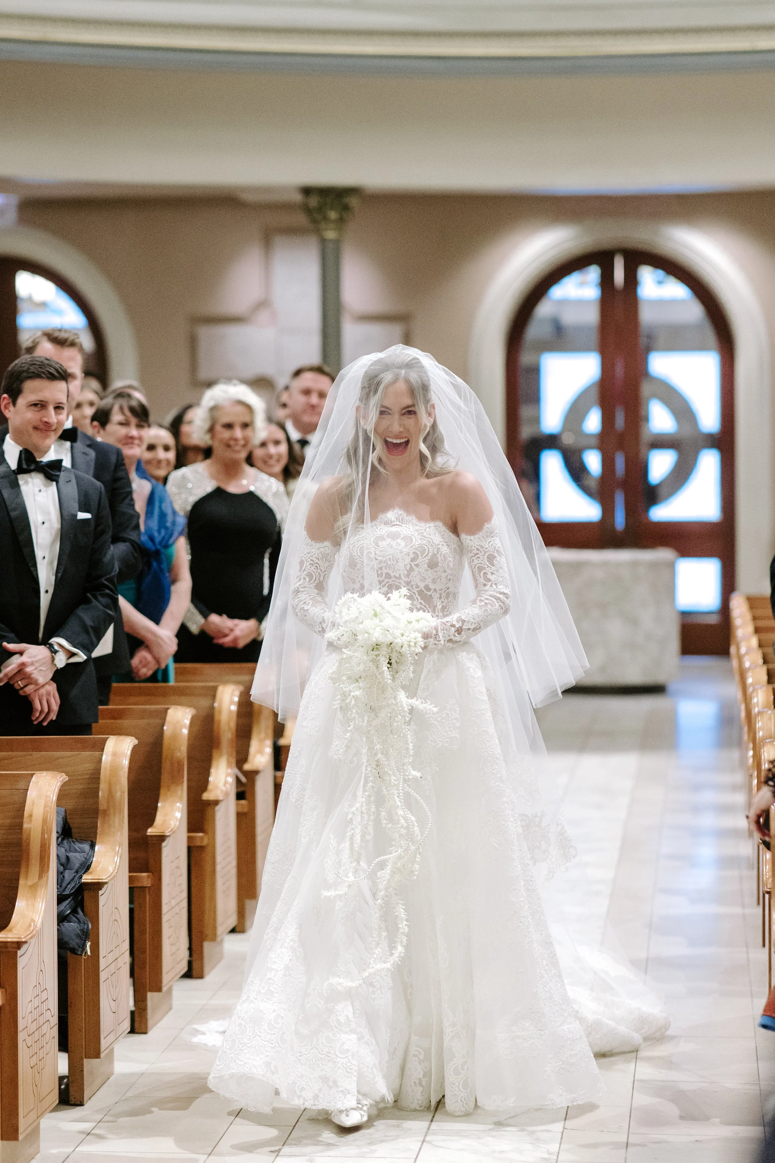 A bride wearing a white lace wedding gown and veil, smiling and holding a bouquet, walking down the aisle in a church during her wedding ceremony.