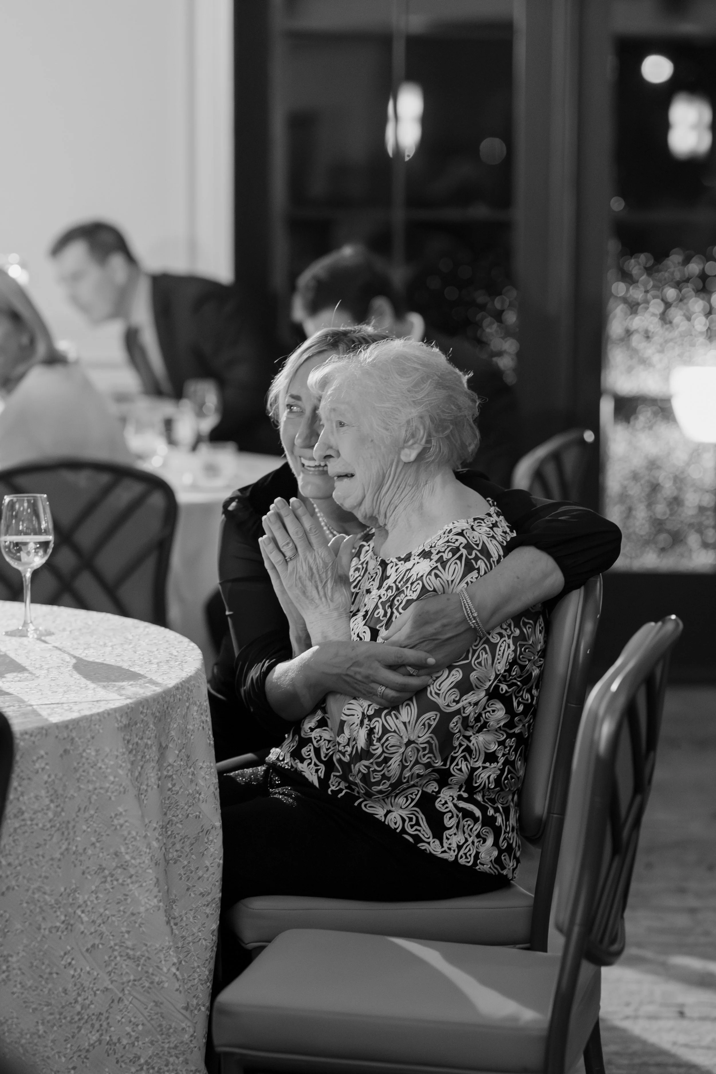 Two women sharing a warm embrace at a formal event, possibly a wedding or celebration, seated at a round table with a patterned tablecloth and glasses of wine, in a room with large windows and dark furniture.