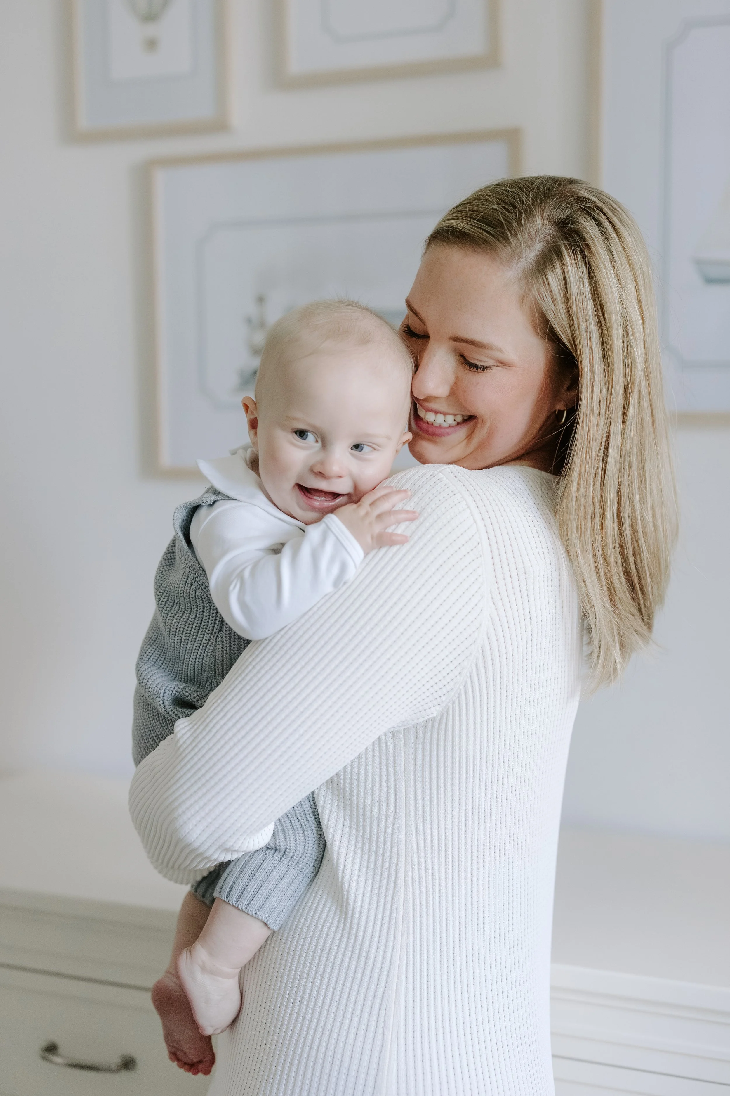 A smiling woman holding a happy baby boy in her arms indoors, with a white dresser and framed pictures on the wall in the background.