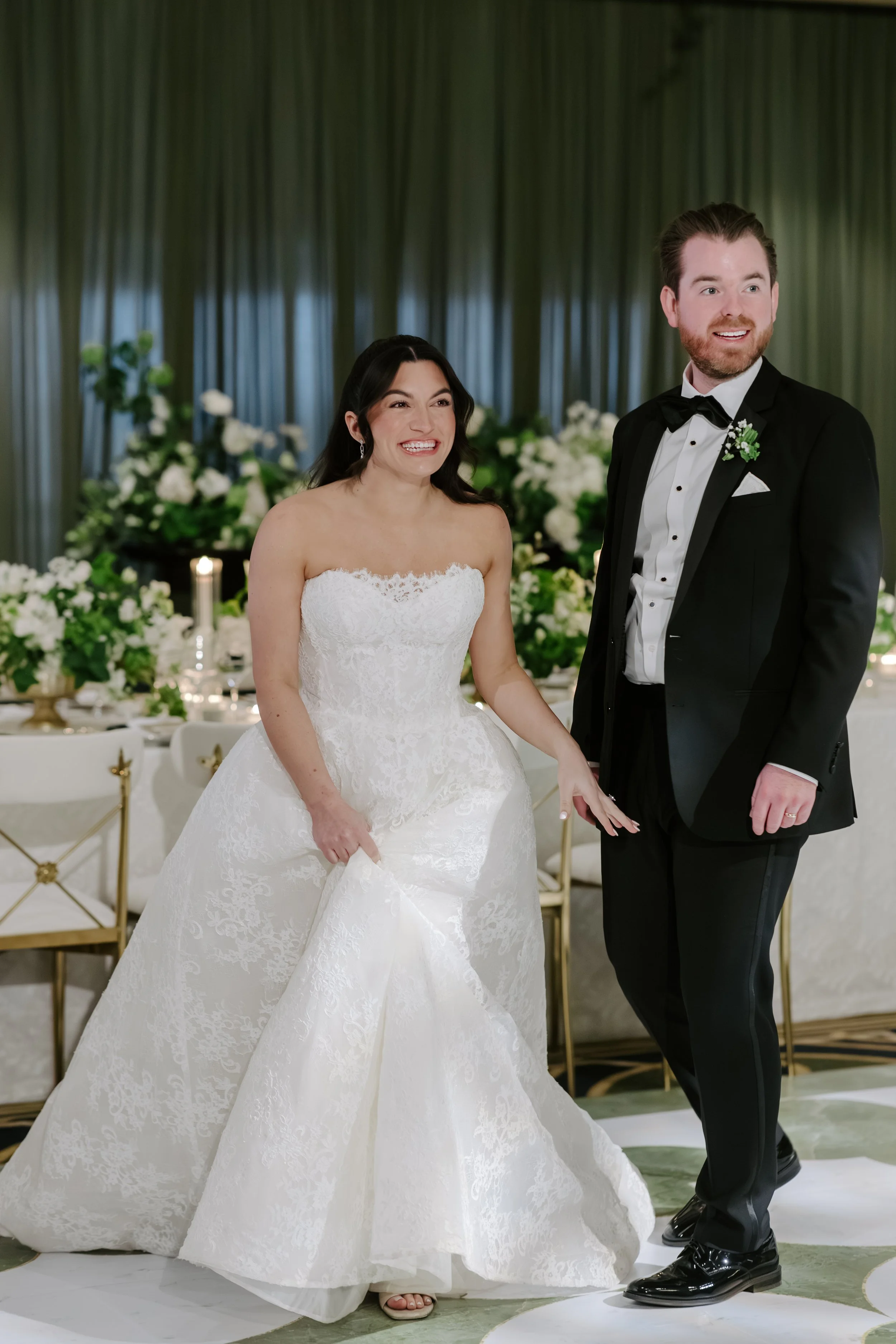 A bride and groom standing together at their wedding reception. The bride is smiling, wearing a white strapless wedding gown, and the groom is also smiling, dressed in a black tuxedo with a bow tie. They are in a decorated venue with floral arrangeme