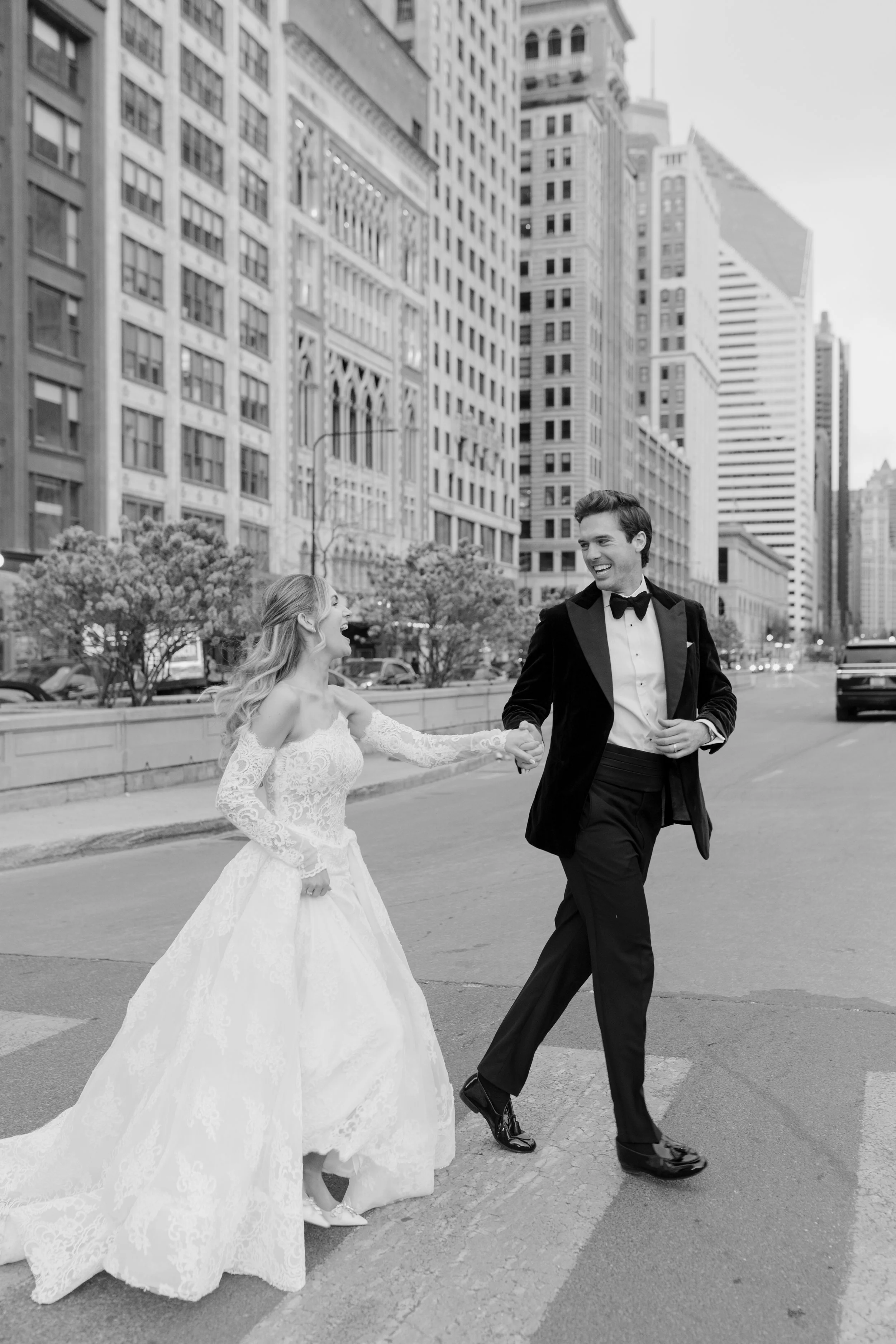 Black and white photo of a couple in wedding attire holding hands and smiling as they walk on a city street with tall buildings in the background.