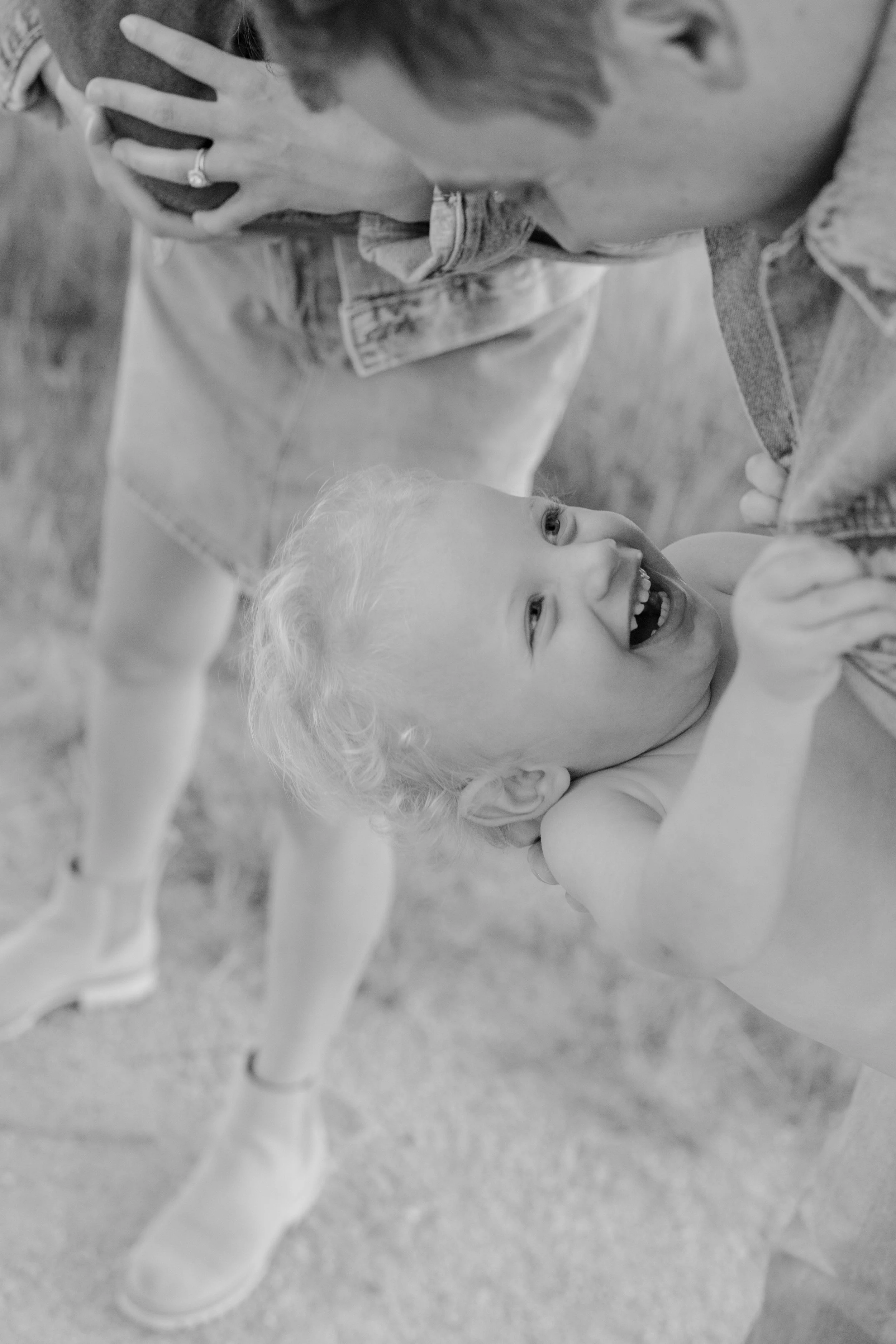 A young child with light curly hair lying on the ground, smiling with mouth open, showing teeth. An adult with glasses holding the child's face, wearing a denim jacket, possibly playing or giving affection. The scene appears to be indoors or on a sof