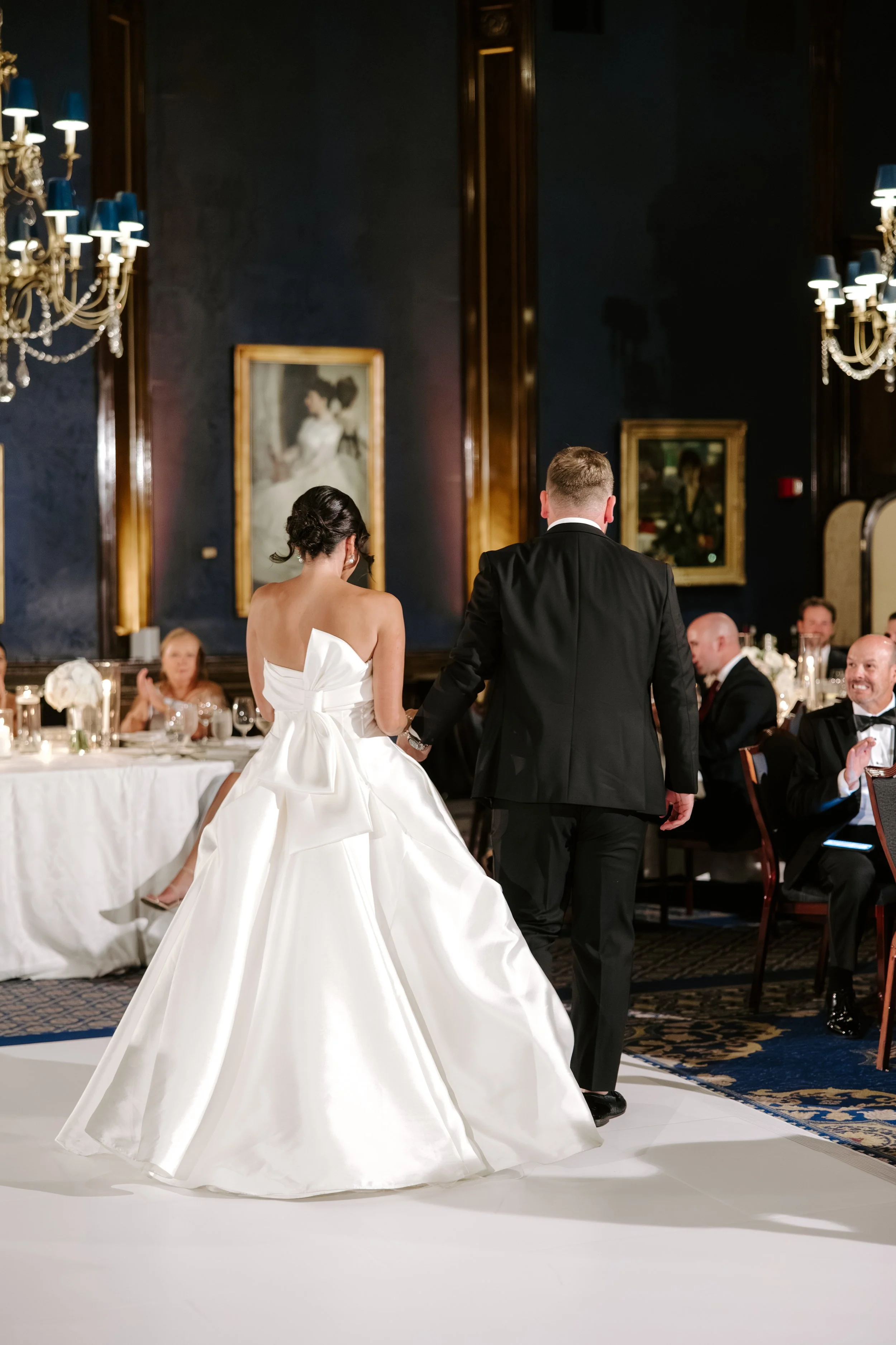 A bride and groom holding hands during their wedding reception in an elegant ballroom with dark walls, chandeliers, and framed artwork.