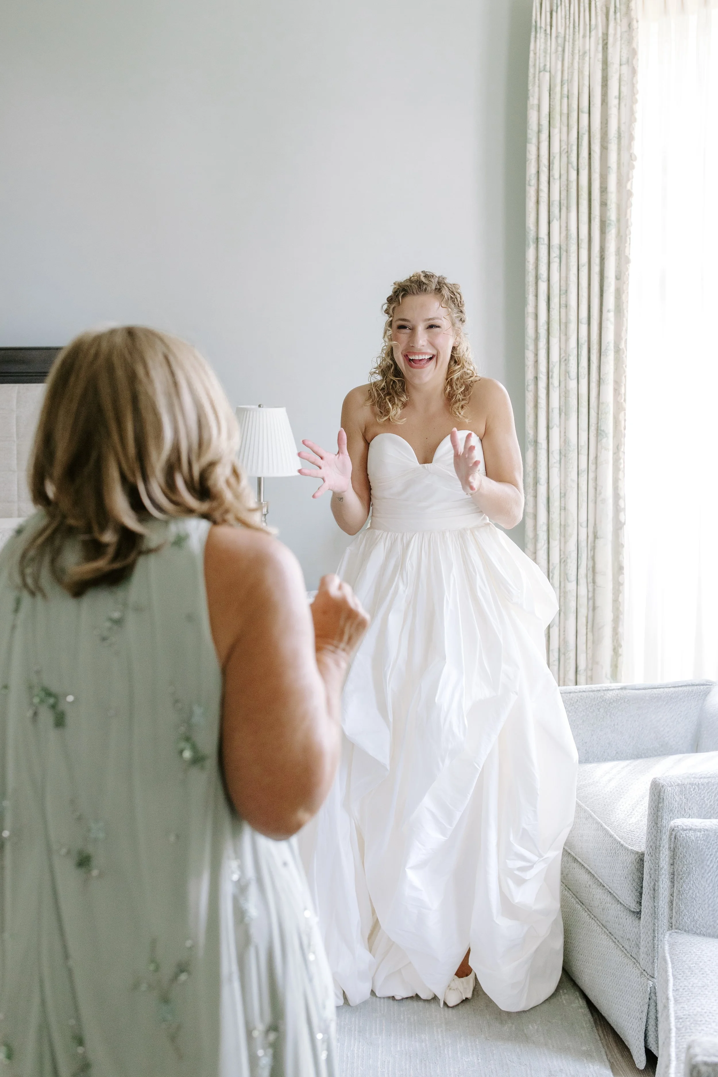 A woman in a white wedding dress is smiling and gesturing while talking to another woman in a light green dress in a well-lit room with a bed, a lamp, and a window with patterned curtains.