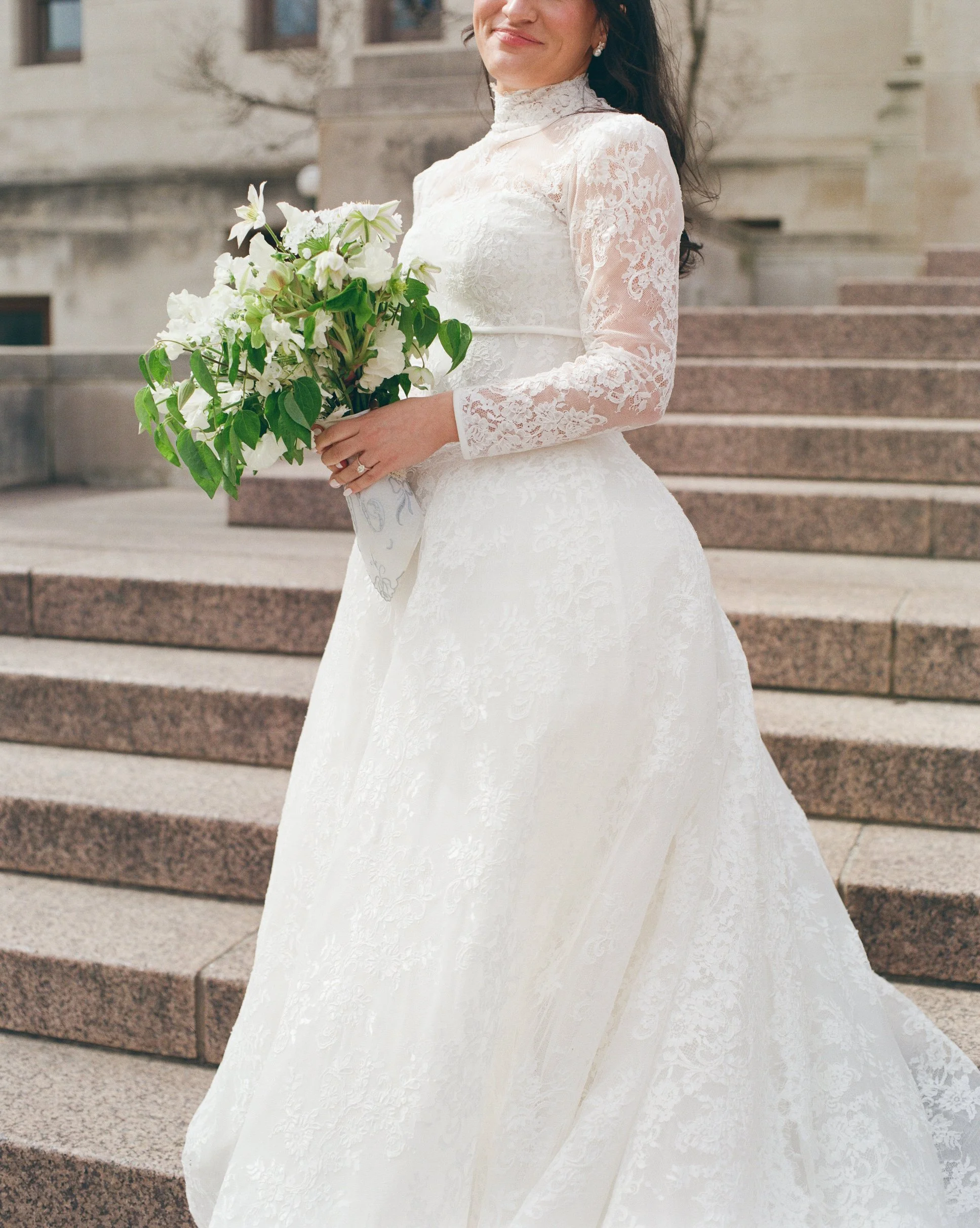 A woman wearing a white lace wedding dress, holding a bouquet of white and green flowers, standing outdoors on stone steps with a building in the background.