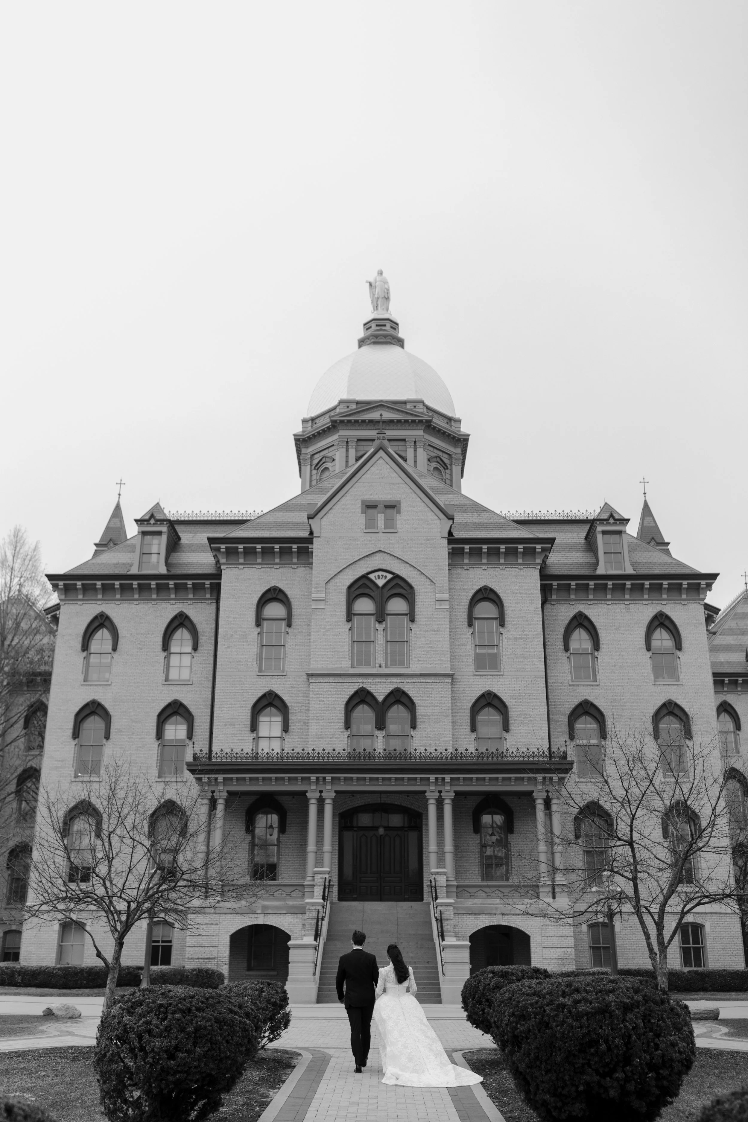 A bride and groom walking towards a large, historic church with a prominent clock tower and a statue on top, black and white photo.
