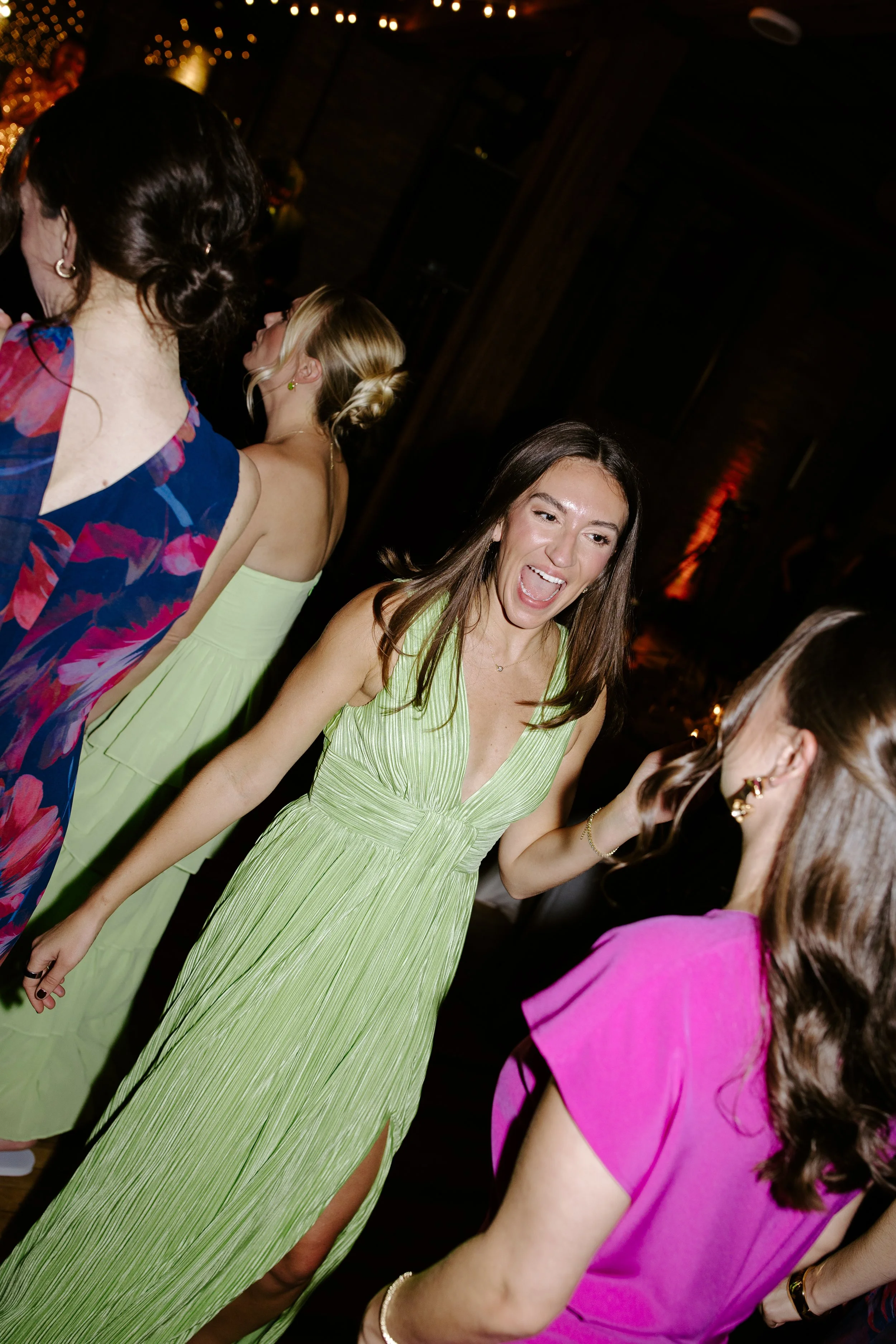 Group of women having fun at a party or celebration, wearing colorful dresses, inside a dimly lit venue with warm lighting and decorative lighting in the background.