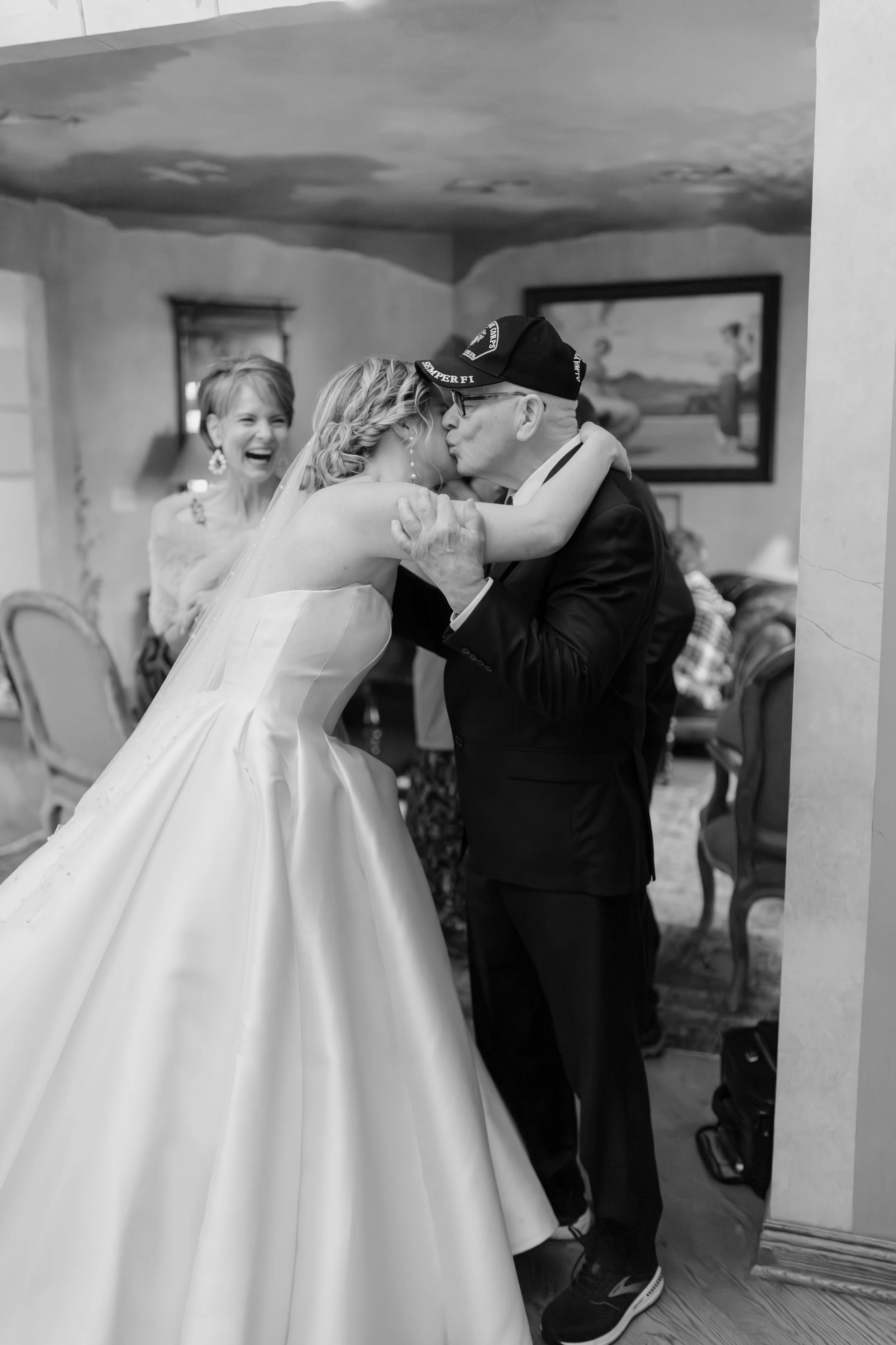 A bride and groom share a kiss during their wedding, with a woman in the background laughing and enjoying the moment.