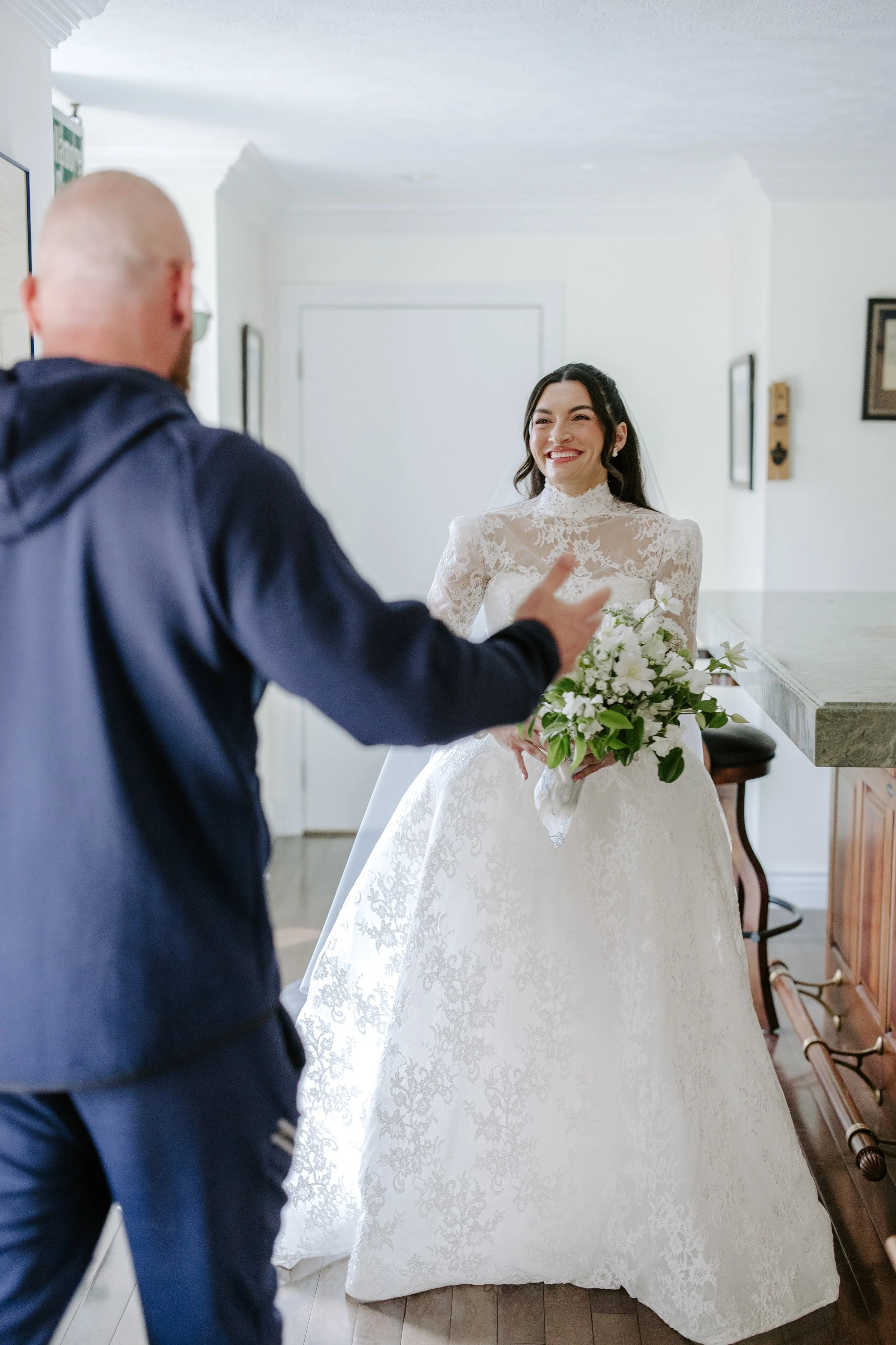 A bride wearing a white wedding dress and holding a bouquet of white flowers smiles as she talks to a man in a blue jacket in an indoor setting with white walls and hardwood floors.