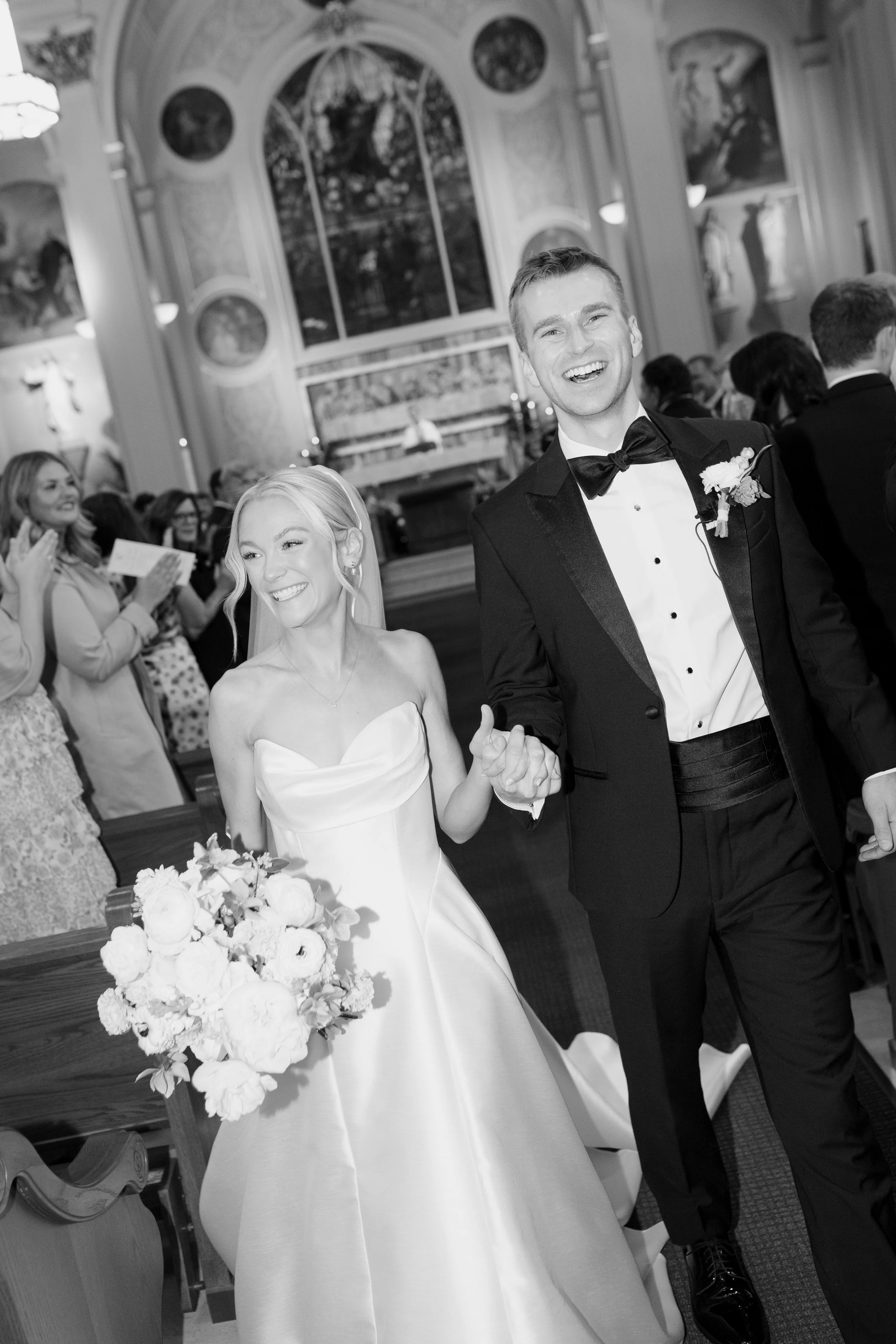 A black and white photo of a wedding ceremony. The bride is holding a bouquet and wearing a strapless wedding gown. The groom is wearing a tuxedo with a bow tie and boutonniere, and they are holding hands. Guests are seated in the background in an or
