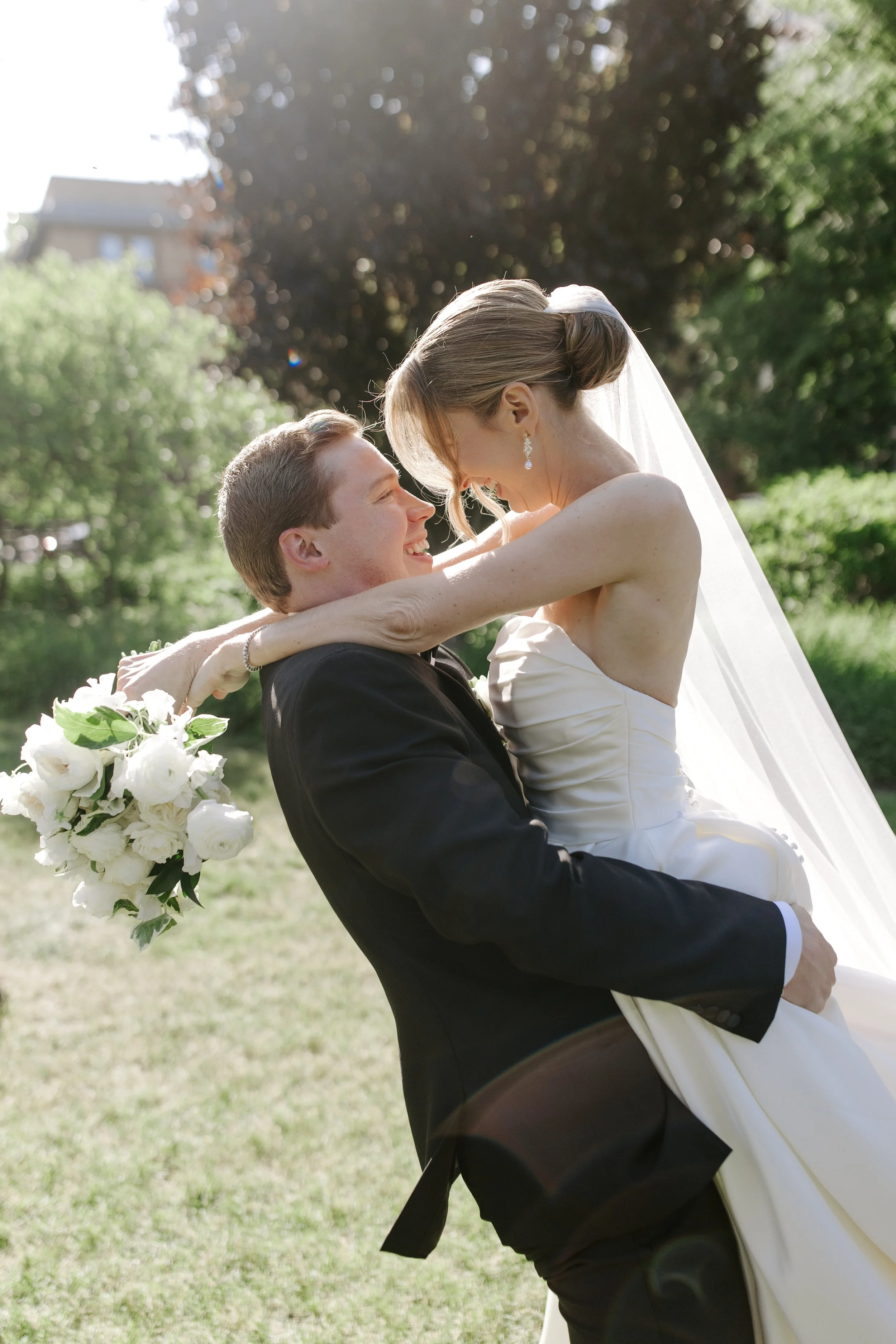 A bride and groom outdoors in a sunny garden, with the groom lifting the bride who holds a bouquet of white flowers, smiling at each other.