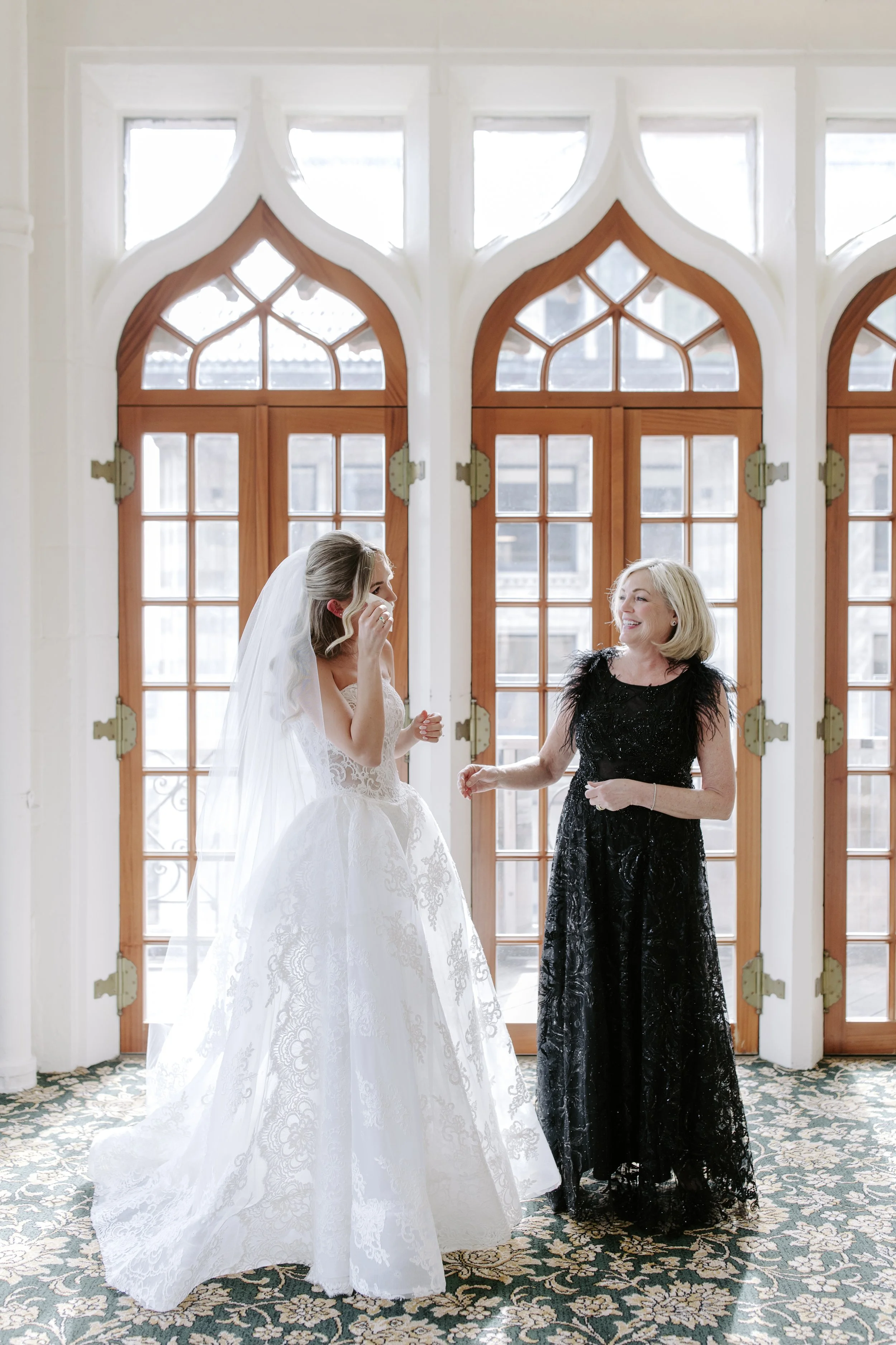 A bride in a white lace wedding dress and veil laughs with an older woman in a black dress in front of large arched windows with wooden frames.