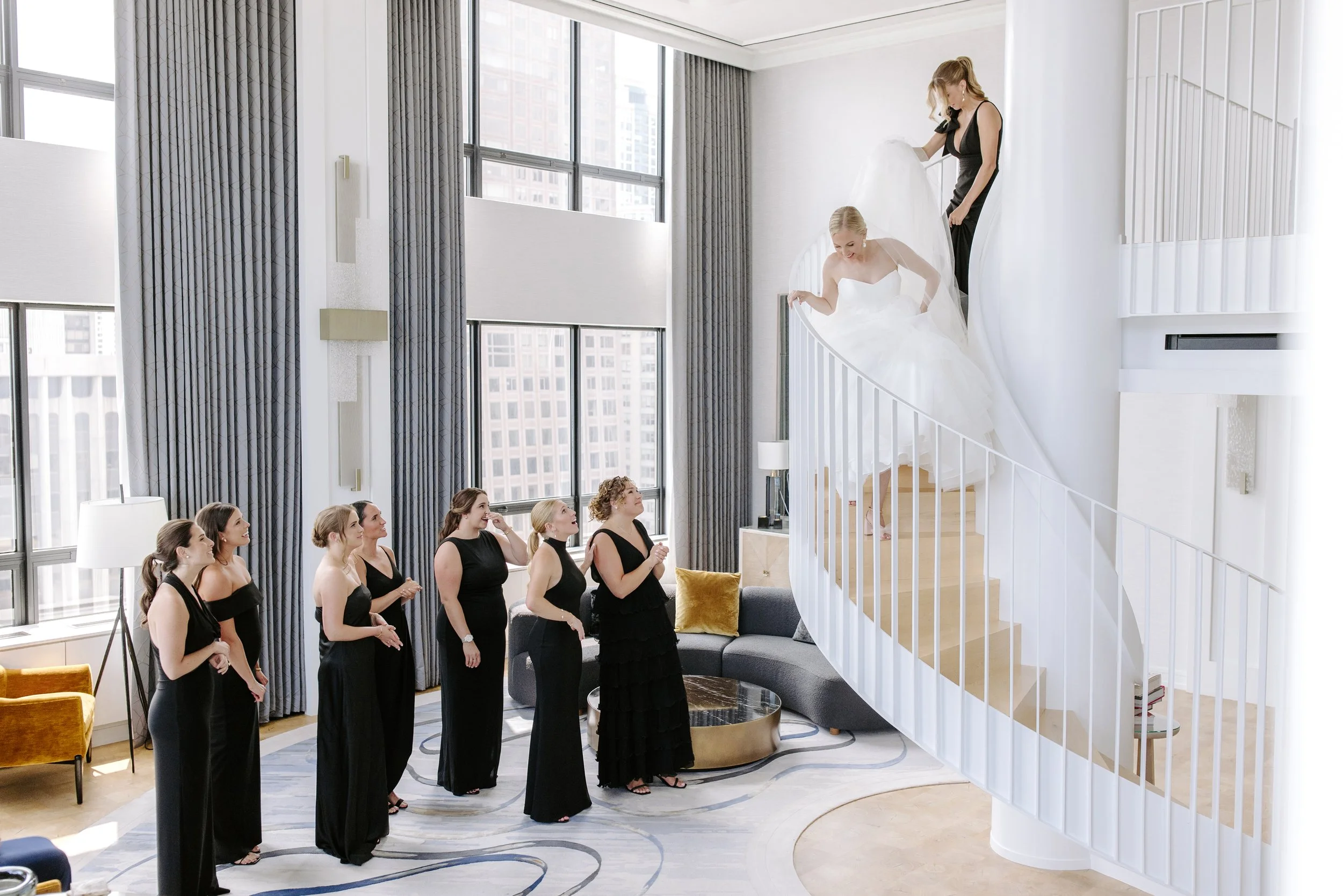 A bride in a white wedding dress descending a staircase with the help of a woman in black, while several women in black dresses look on and smile in a bright, modern living room with large windows and city views.