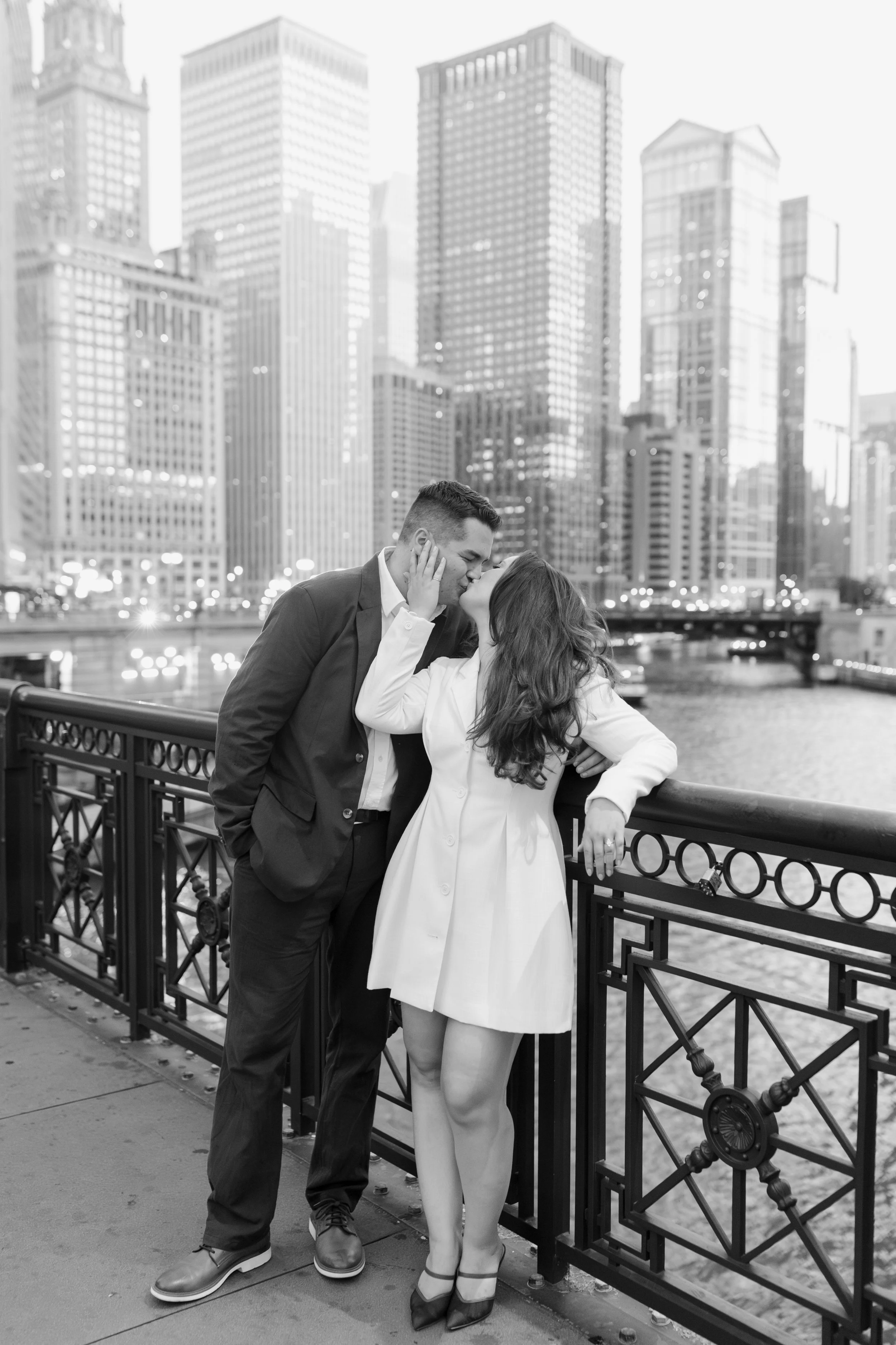 A black and white photo of a couple kissing on a bridge with city skyscrapers in the background, a river below, and a metal railing.