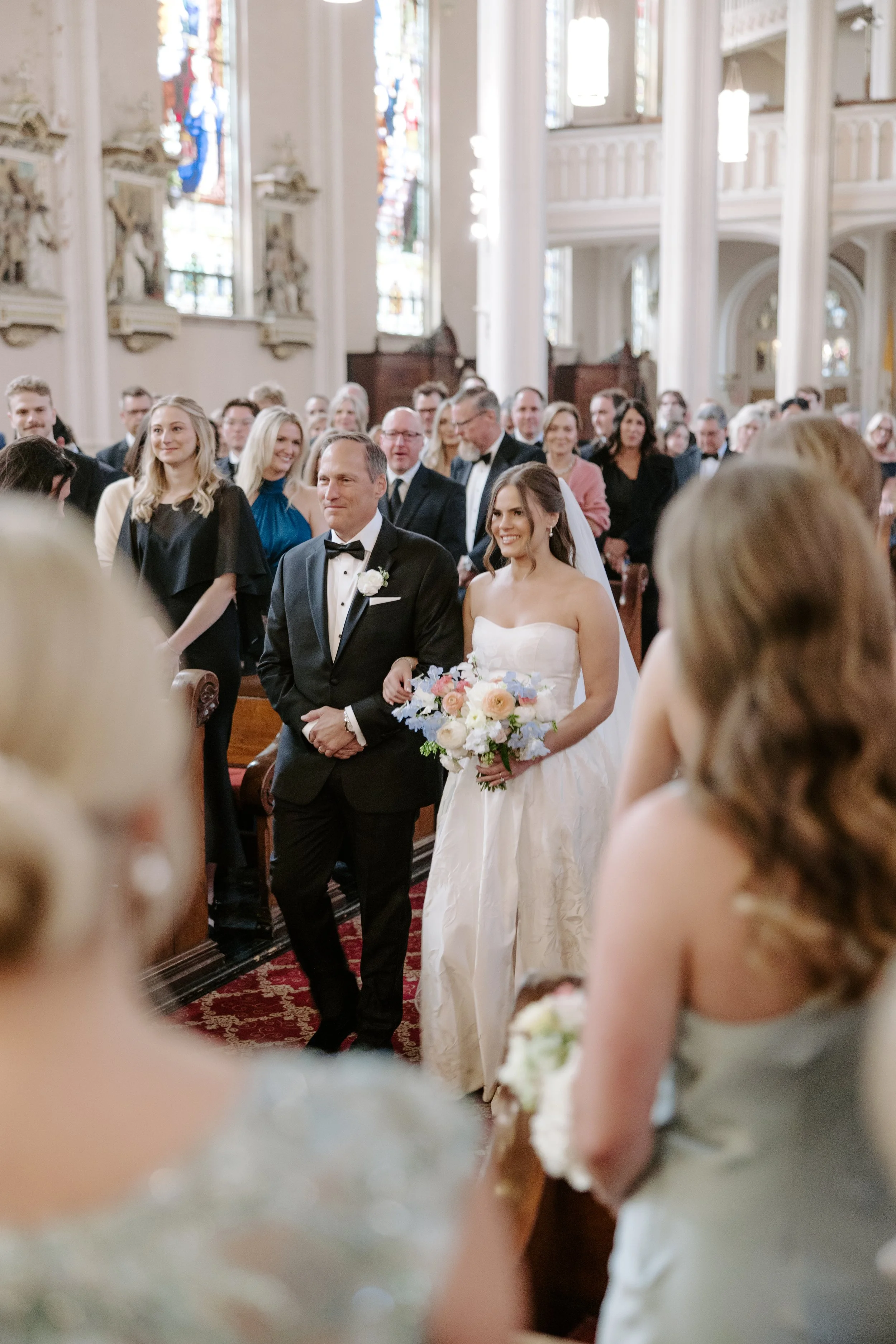 A bride in a white wedding dress holding a bouquet walking down the aisle with her father during a wedding ceremony inside a church with high ceilings, stained glass windows, and guests seated on either side.