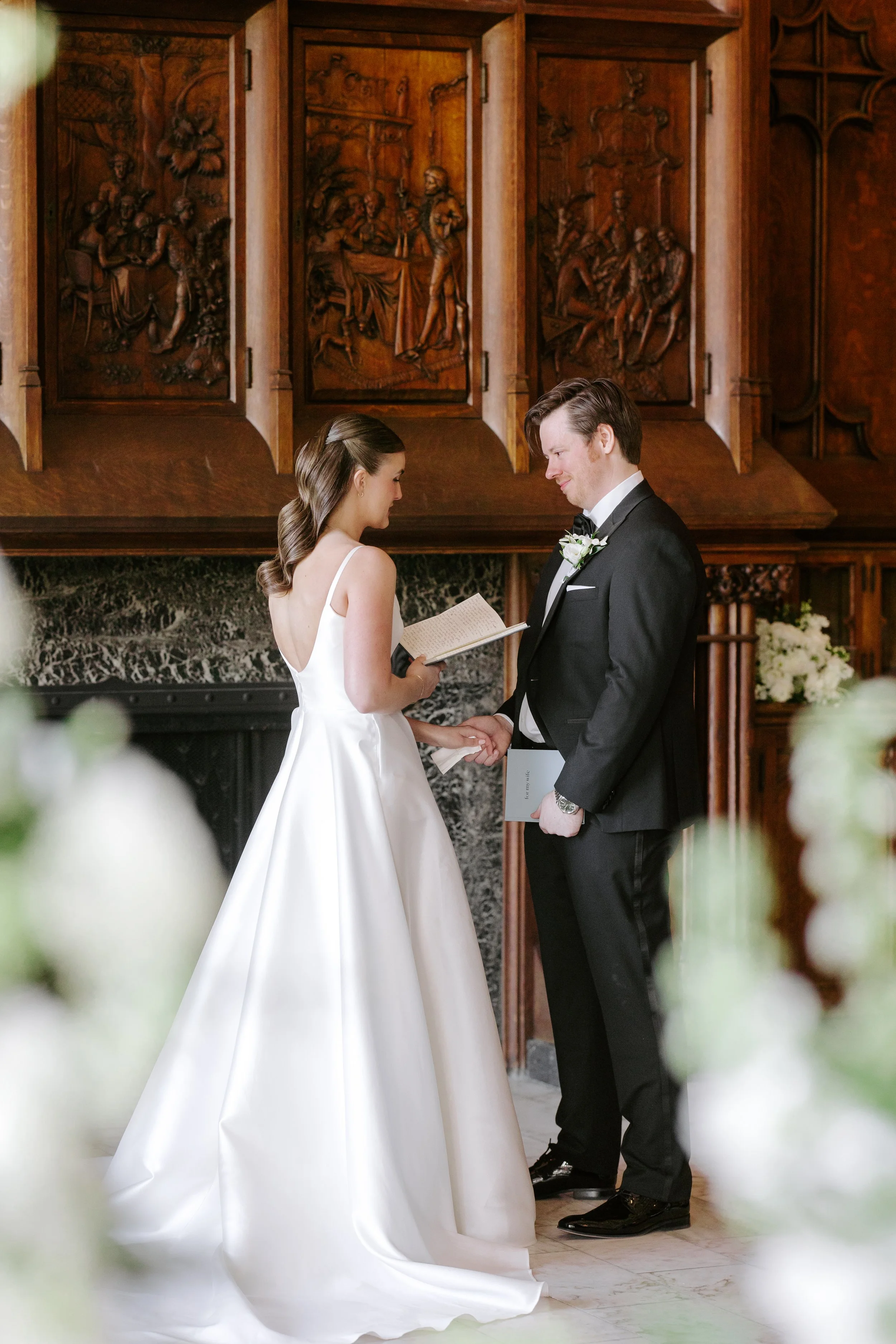 A bride and groom holding hands and exchanging vows during a wedding ceremony inside a room with wooden paneling featuring carved scenes.