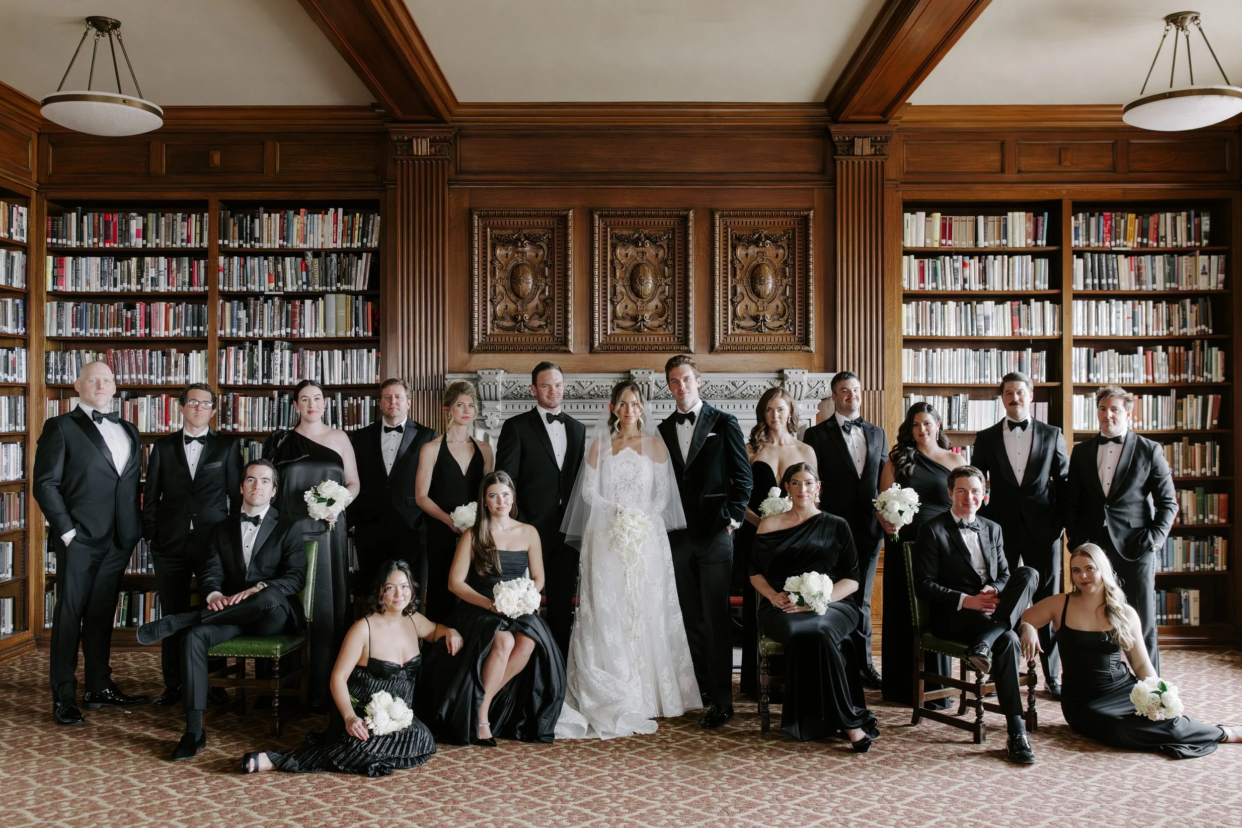 A wedding party of men and women in formal attire, with the bride in a white wedding gown and the groom in a black tuxedo, standing in a library with tall wooden bookshelves and ornate wood paneling.