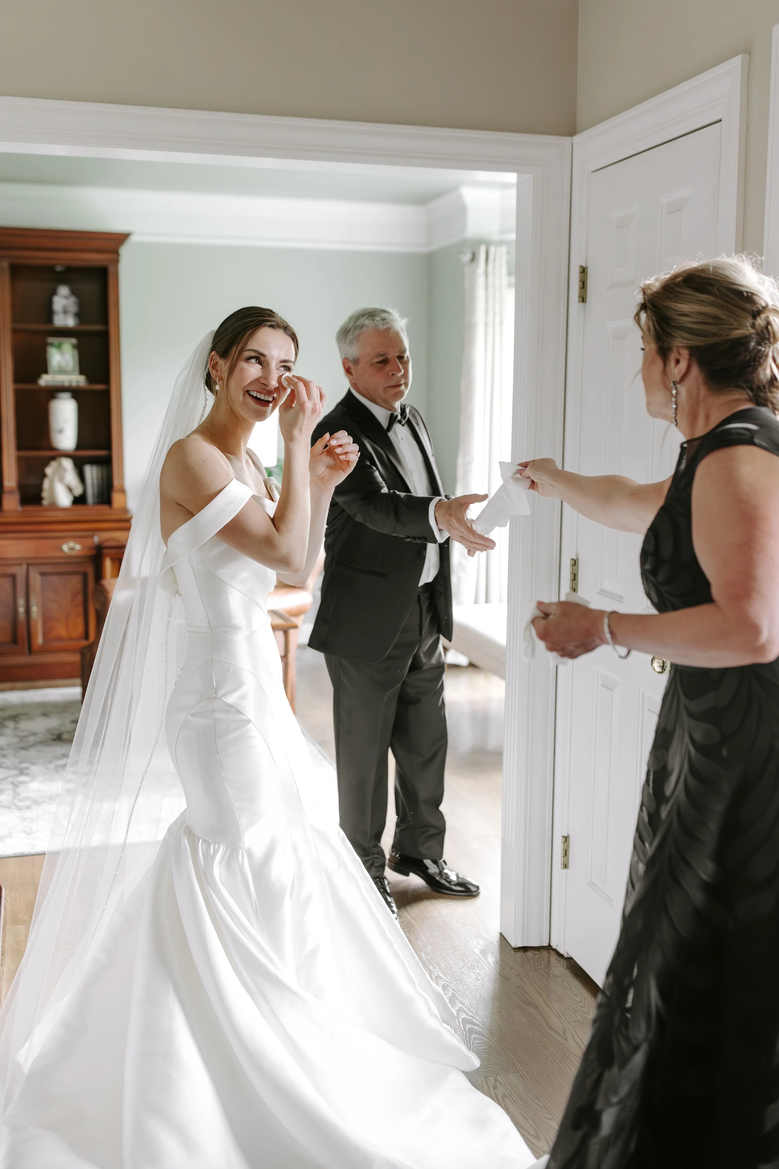 A bride in a white wedding dress is emotional and wiping away tears while standing in a room, and a man in a tuxedo is giving her a tissue. A woman in a black dress extends her hand holding the tissue towards the bride.