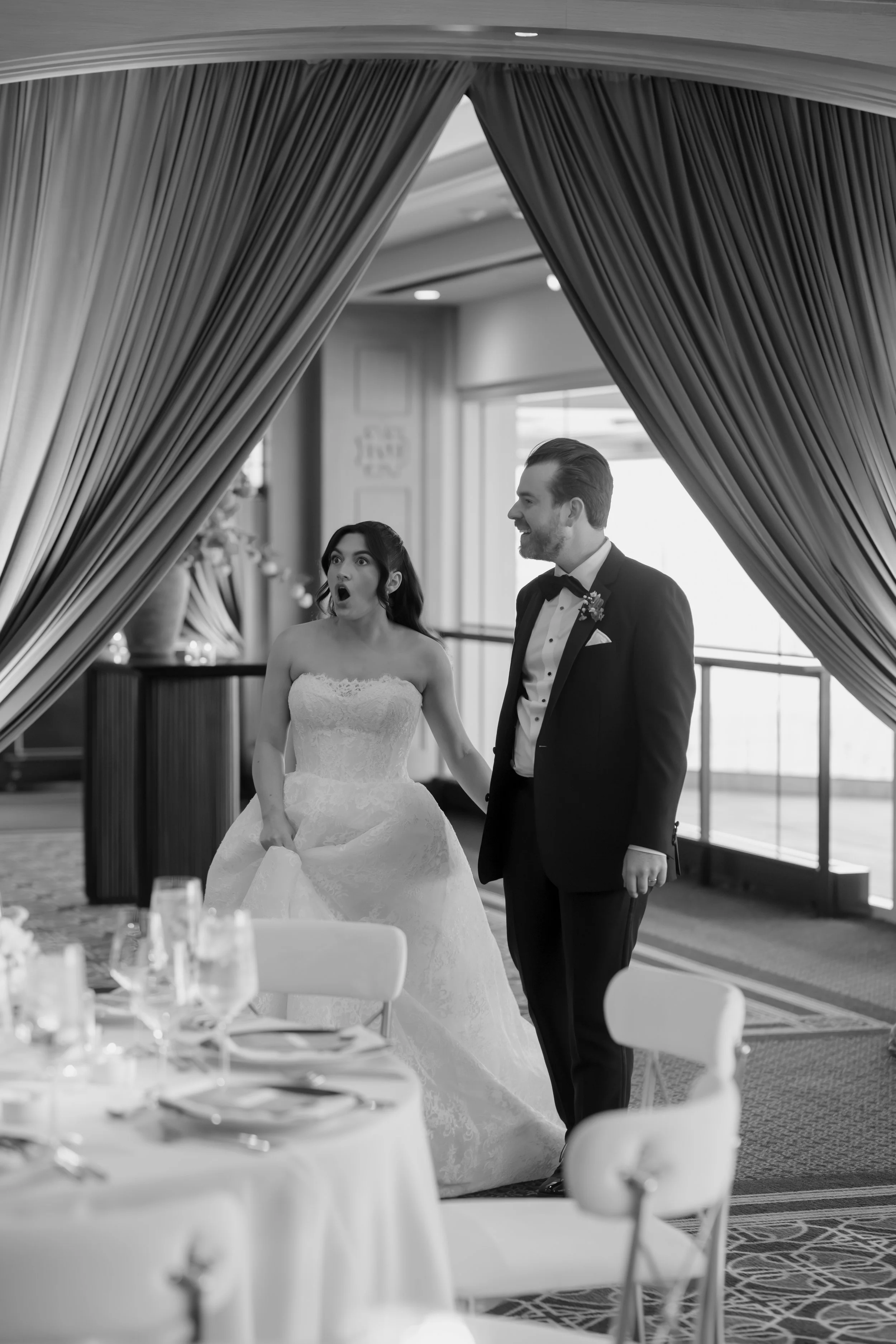 A bride and groom in formal attire standing together at a wedding reception, with the bride appearing surprised and the groom smiling.