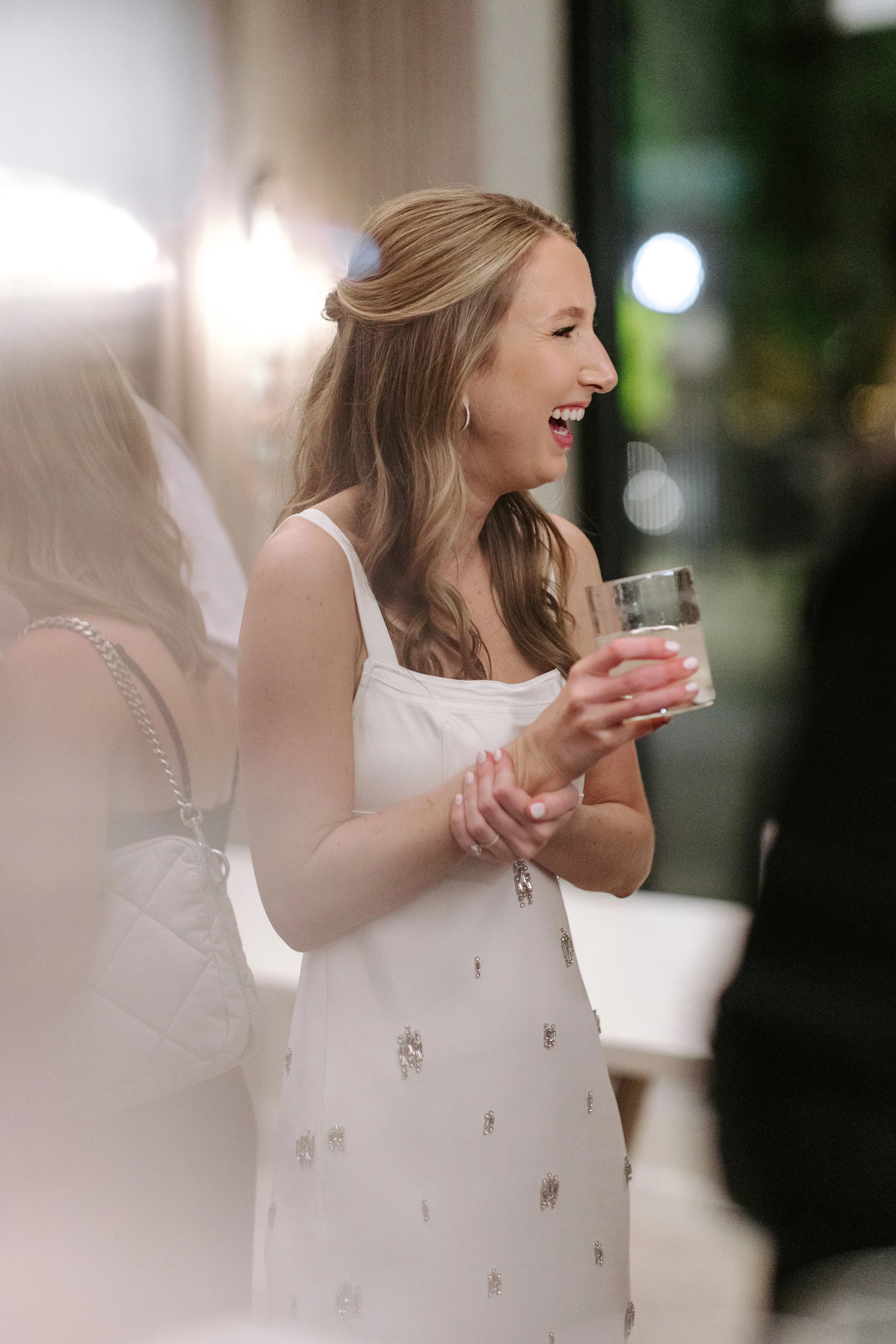 A woman with long light brown hair wearing a white dress with decorative silver embellishments, holding a glass of water, and smiling while looking to the side at an indoor social event.